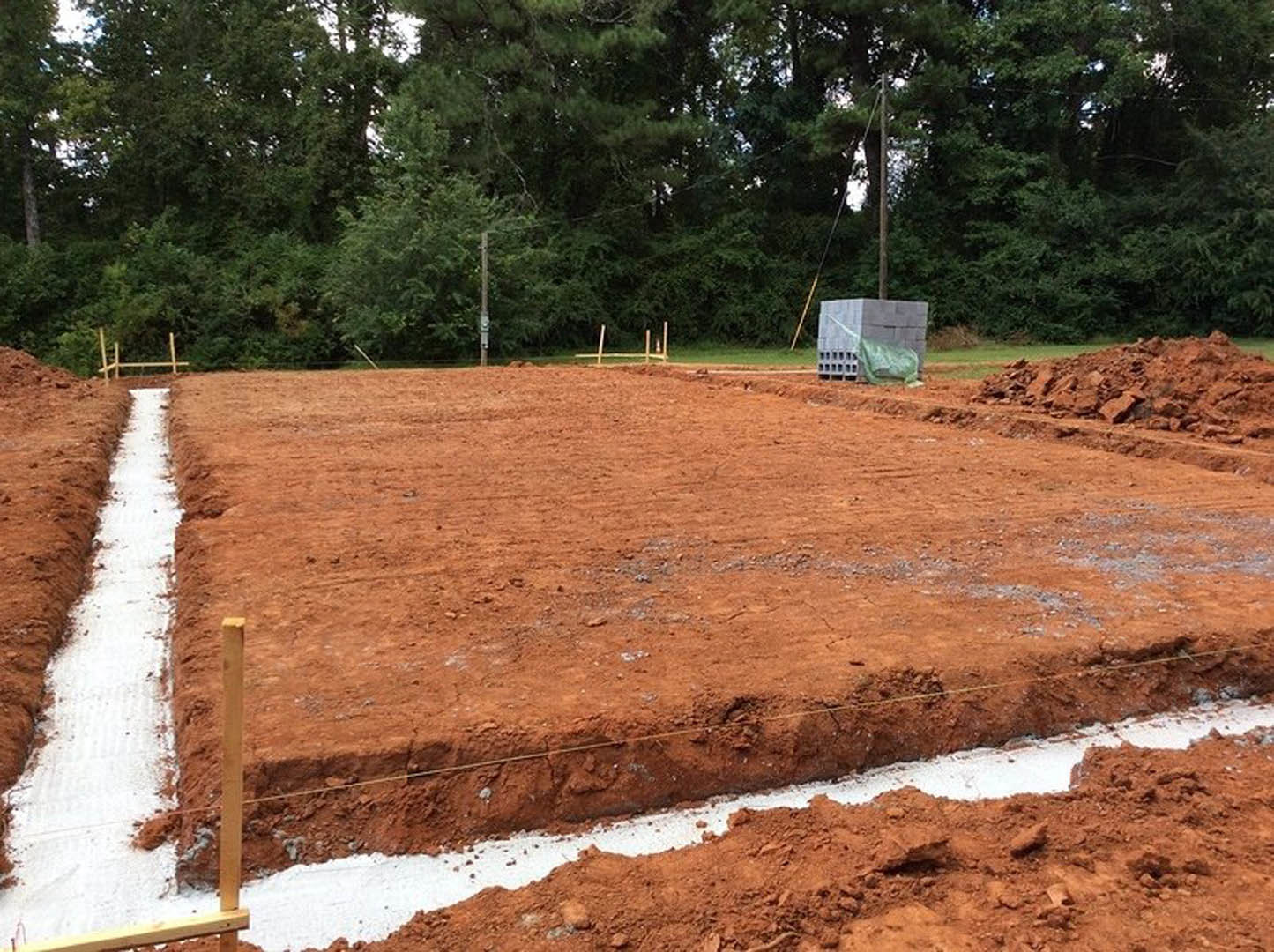 Dirt construction site bordered by trees, stacked bricks on pallets, wire fencing, snow-covered path in a trench