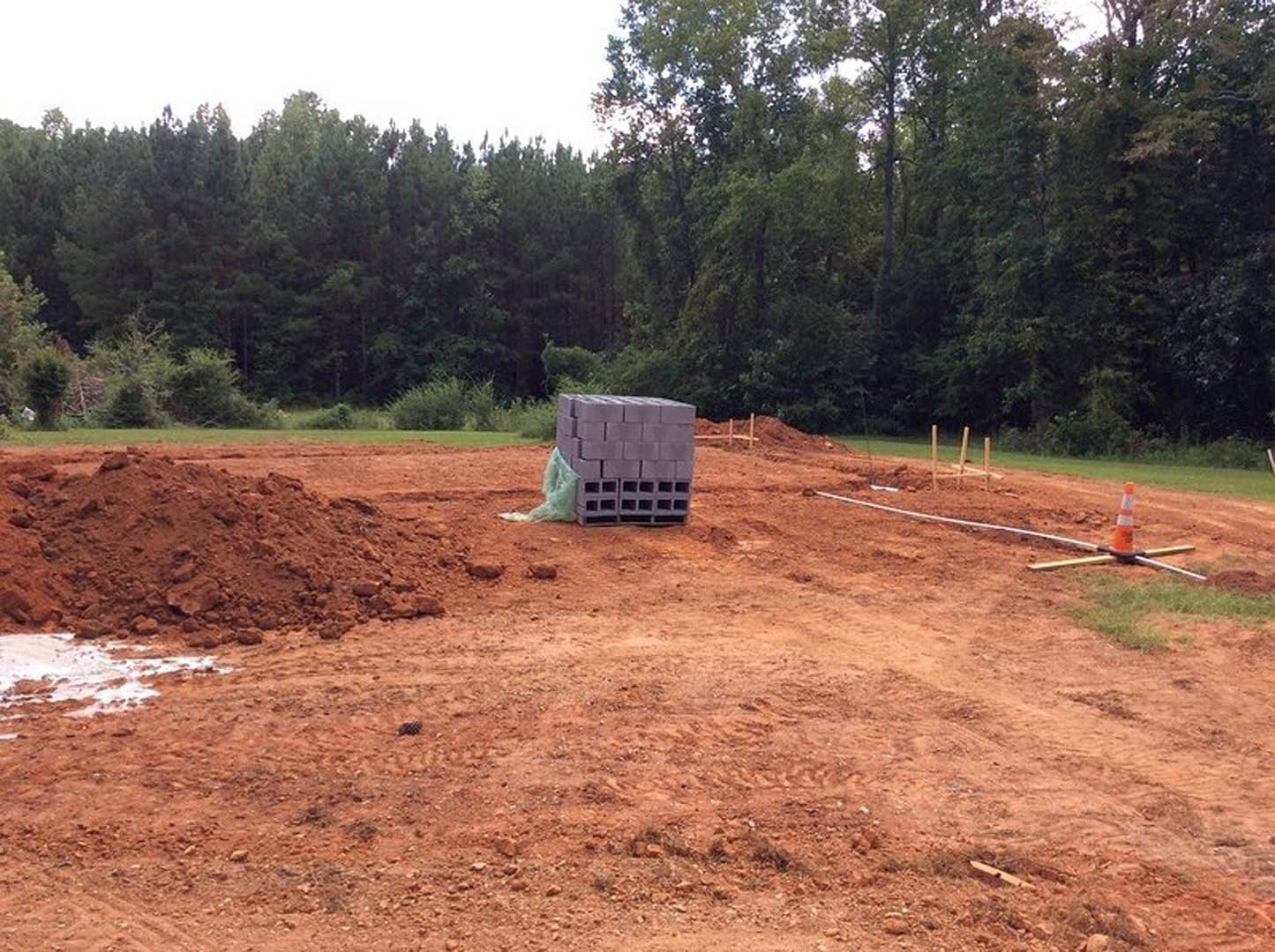 Stack of grey bricks and mound of dirt on muddy construction site with grass, trees, and cloudy sky in background