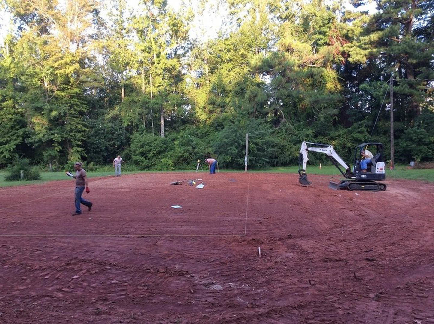 Construction crew preparing dirt foundation on open field bordered by mature trees, with workers handling tools and machinery, scattered equipment visible on soil.