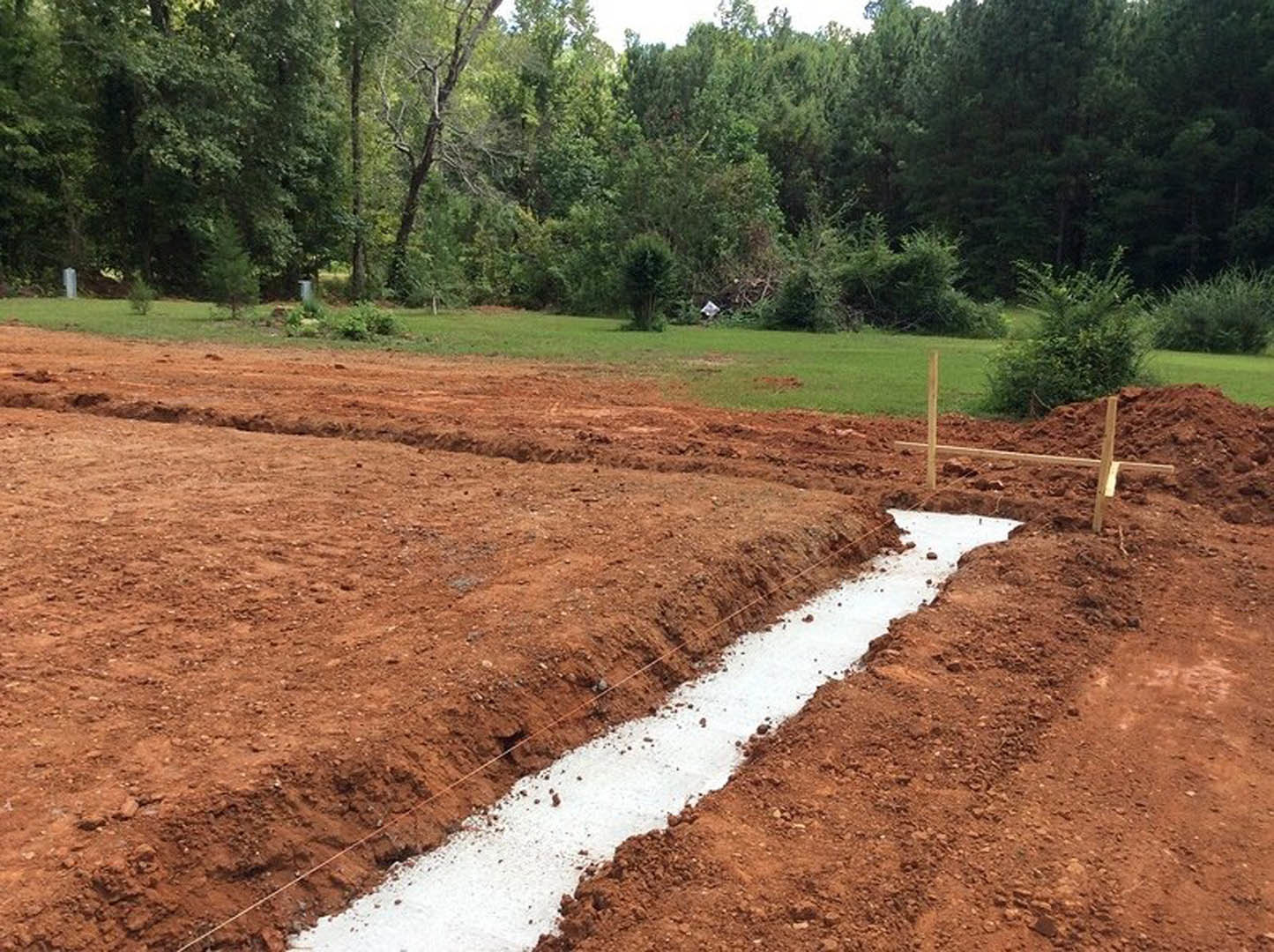 Wooden fence bordering a construction site with a dirt ditch, white marking line, grassy field, trees, and green bush