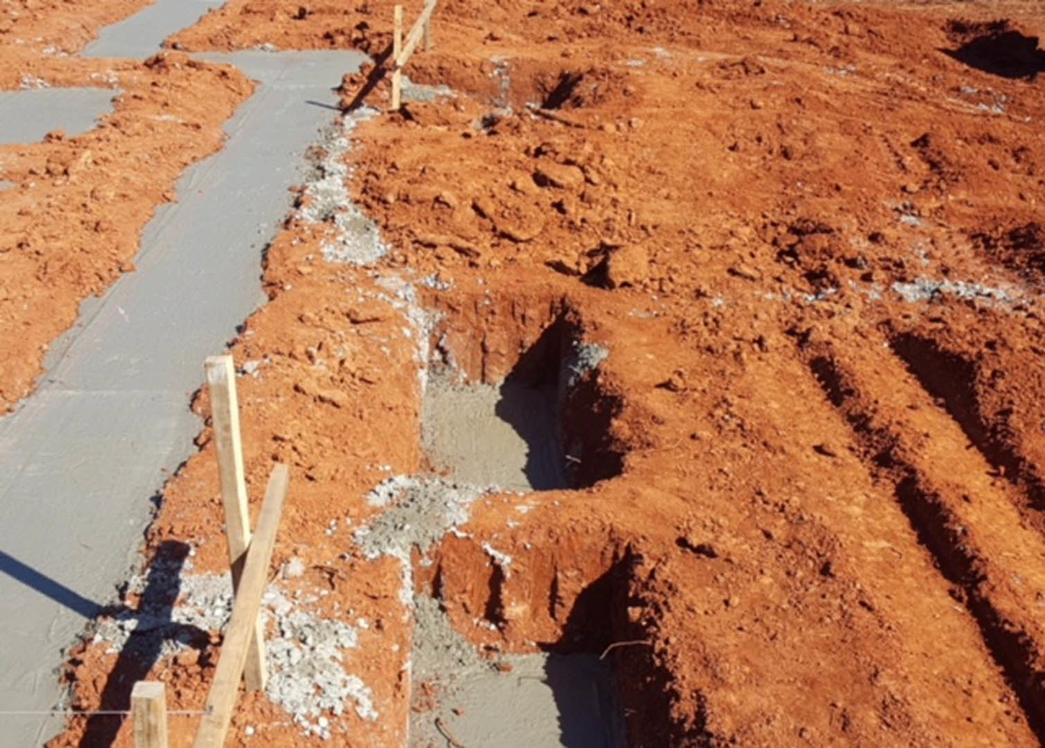 Deep trench dug into brown soil beside a dirt pile, bordered by a wooden fence and metal pole, outdoor residential construction site.