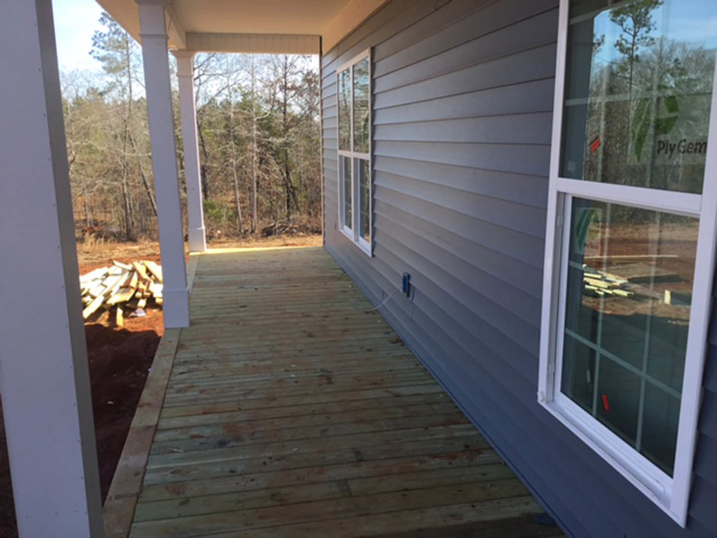 Two-story home with light siding, covered front porch, wood deck, large windows, stacked firewood near entry, mature trees and shrubs in landscaped yard