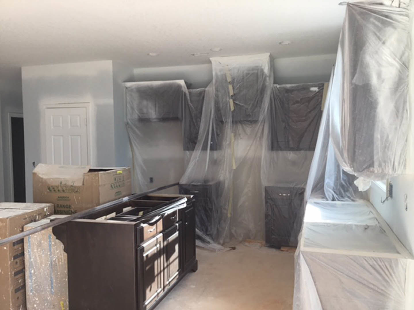 Kitchen with white cabinets, drawers, and boxes on the floor; plastic coverings draped over walls; white door with a sign; unfinished surfaces and scattered materials.
