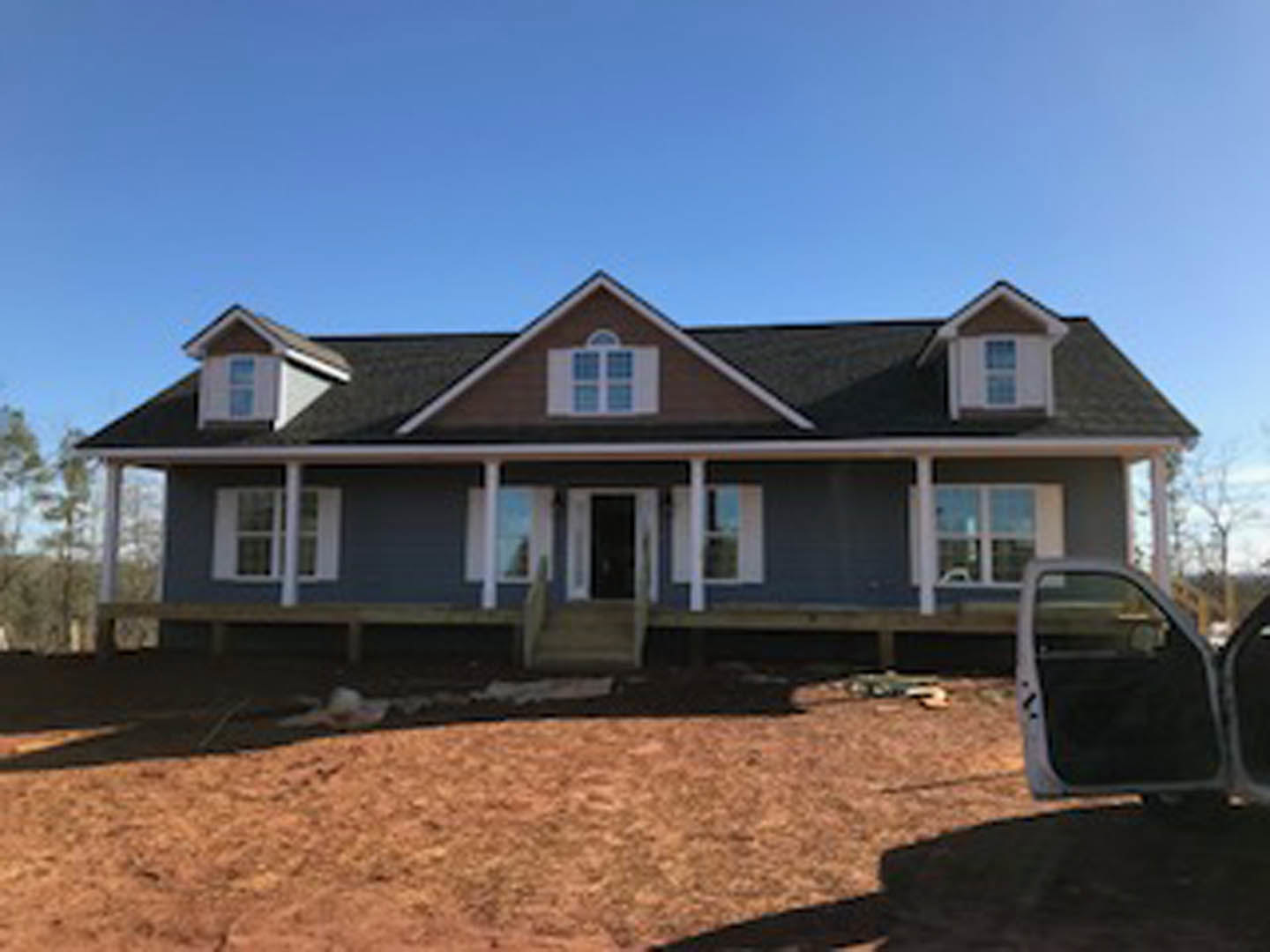 Two-story house under construction with exposed framing, partially finished siding, covered porch with stairs, parked car in driveway, tree and blue sky in background