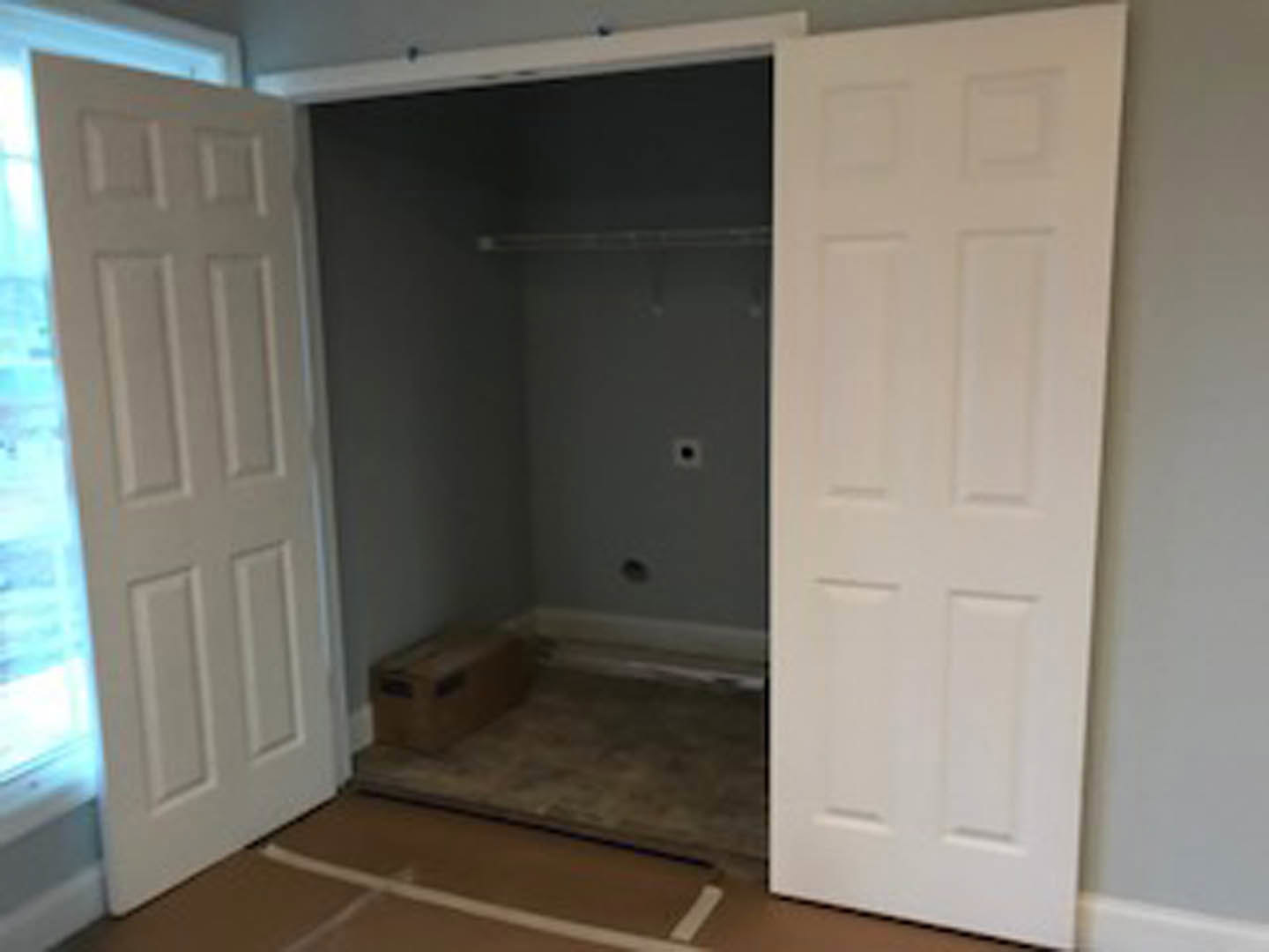 White double closet doors with silver handles set in a light grey wall, open to reveal a brown storage box on wood flooring, with a blurred window in the background.