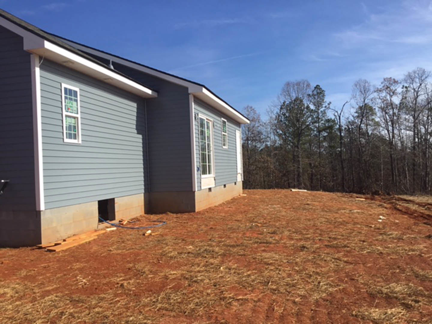 Modern home with light siding, large windows, and blue hose on dirt yard; mature trees and blue sky in background