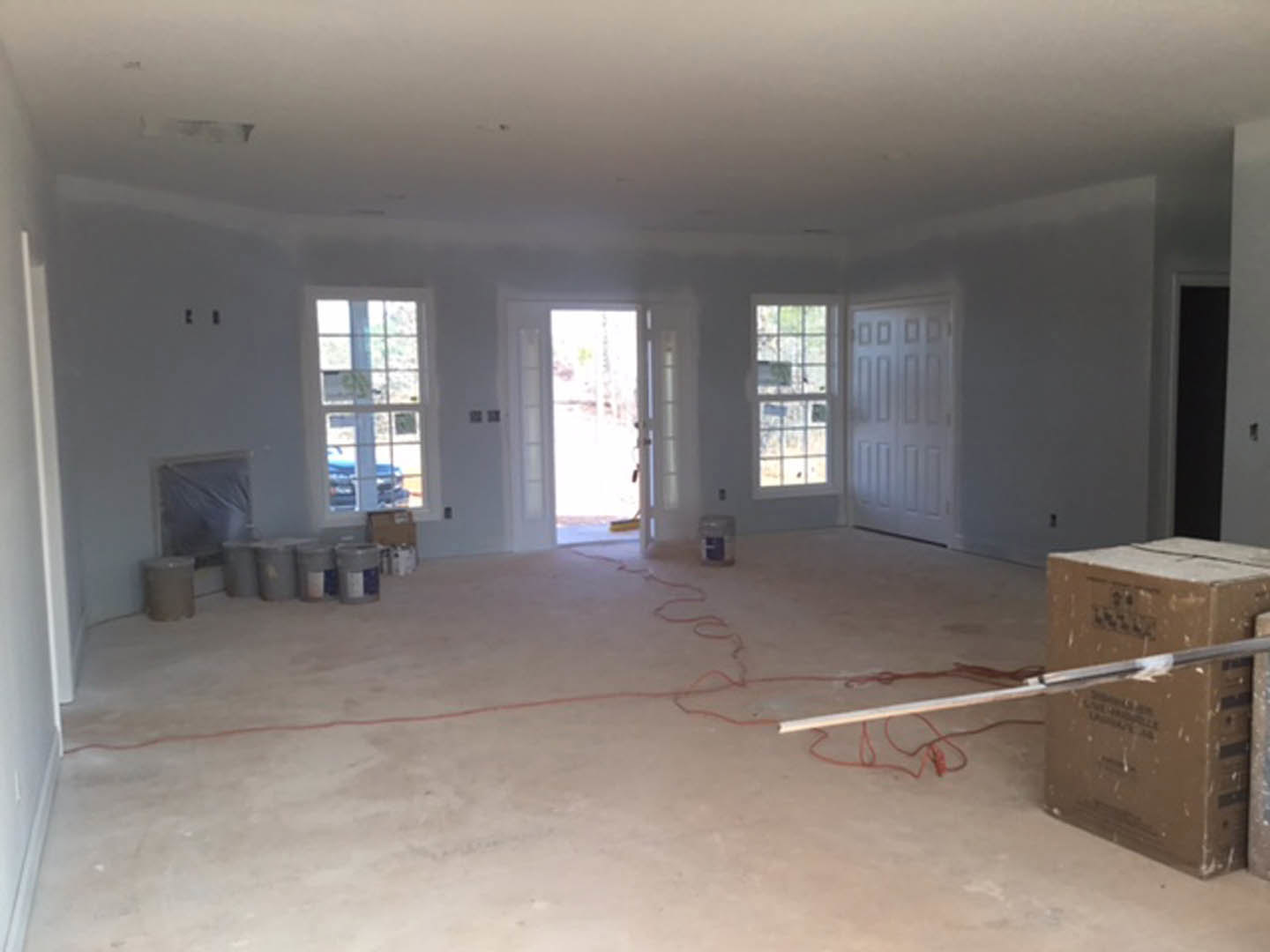 Unfinished room with white plaster walls, paint cans, metal ladder, white door illuminated by natural light, plastic storage container with blue lid, and trash can on bare flooring