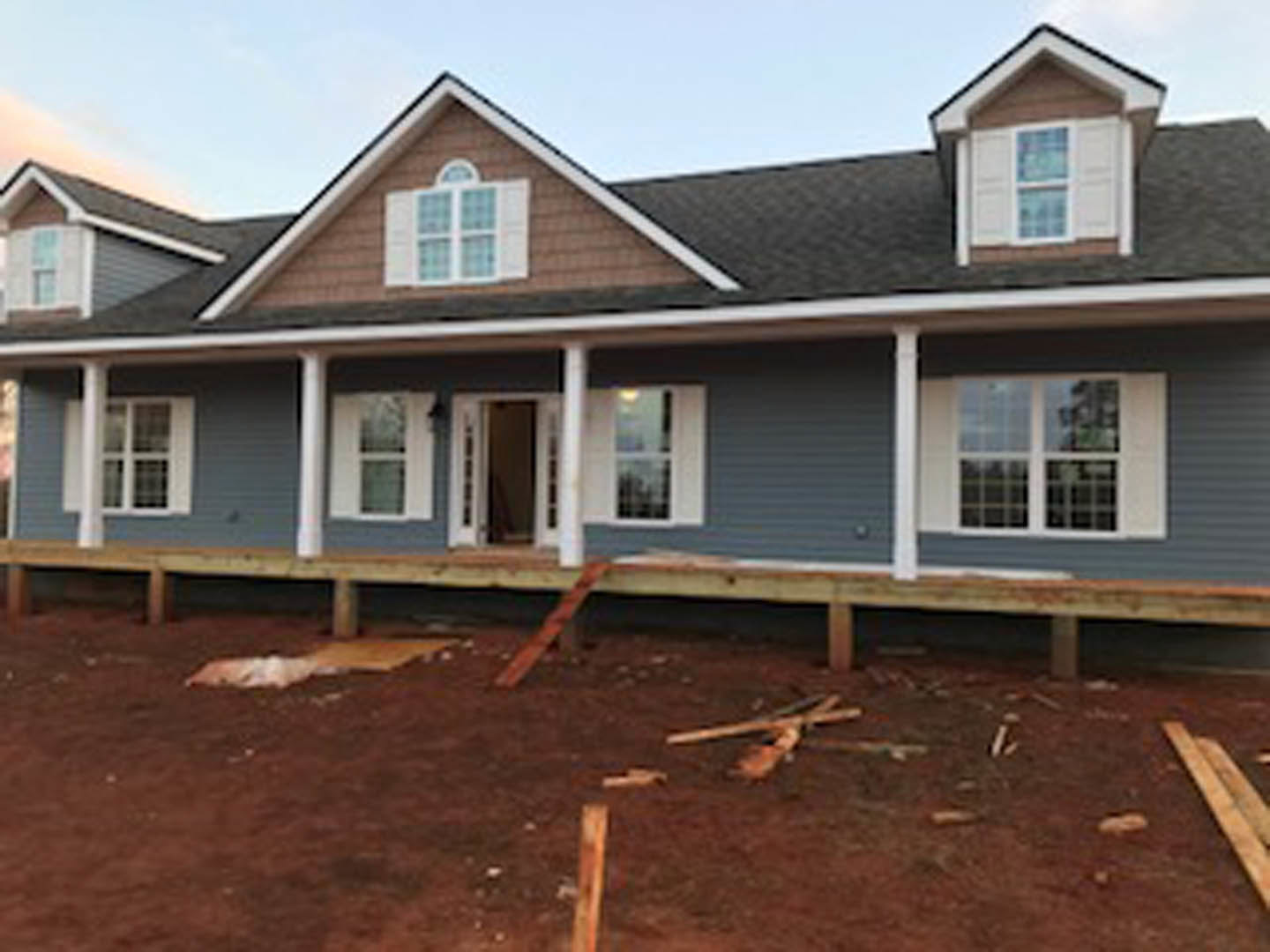 Partially built house with blue roof, white-framed window, exposed wood framing, and dirt yard in foreground