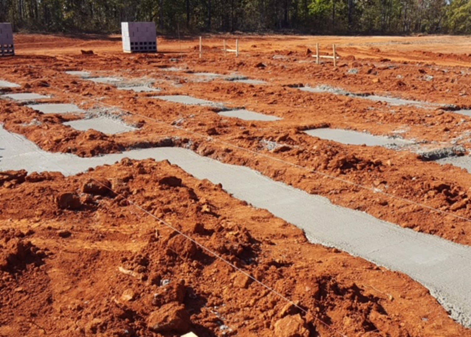Concrete foundation and stacked white blocks on a dirt construction site, with a fence and trees in the background