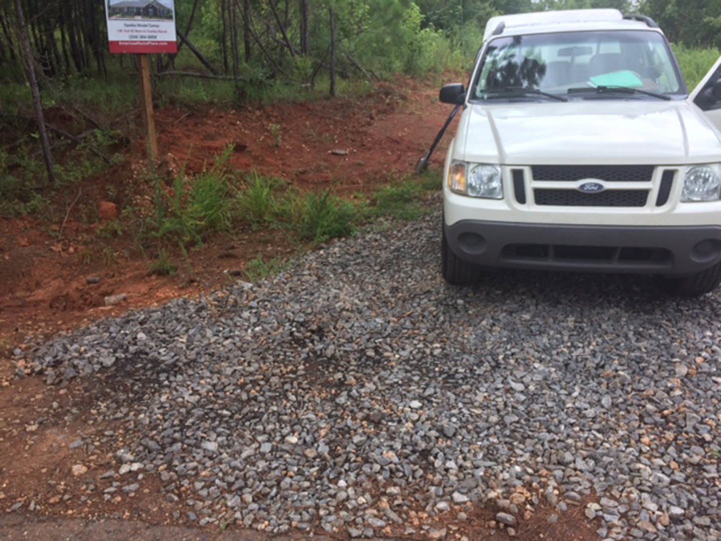White pickup truck with open doors parked on gravel driveway beside a wooden sign, surrounded by trees and forested landscape