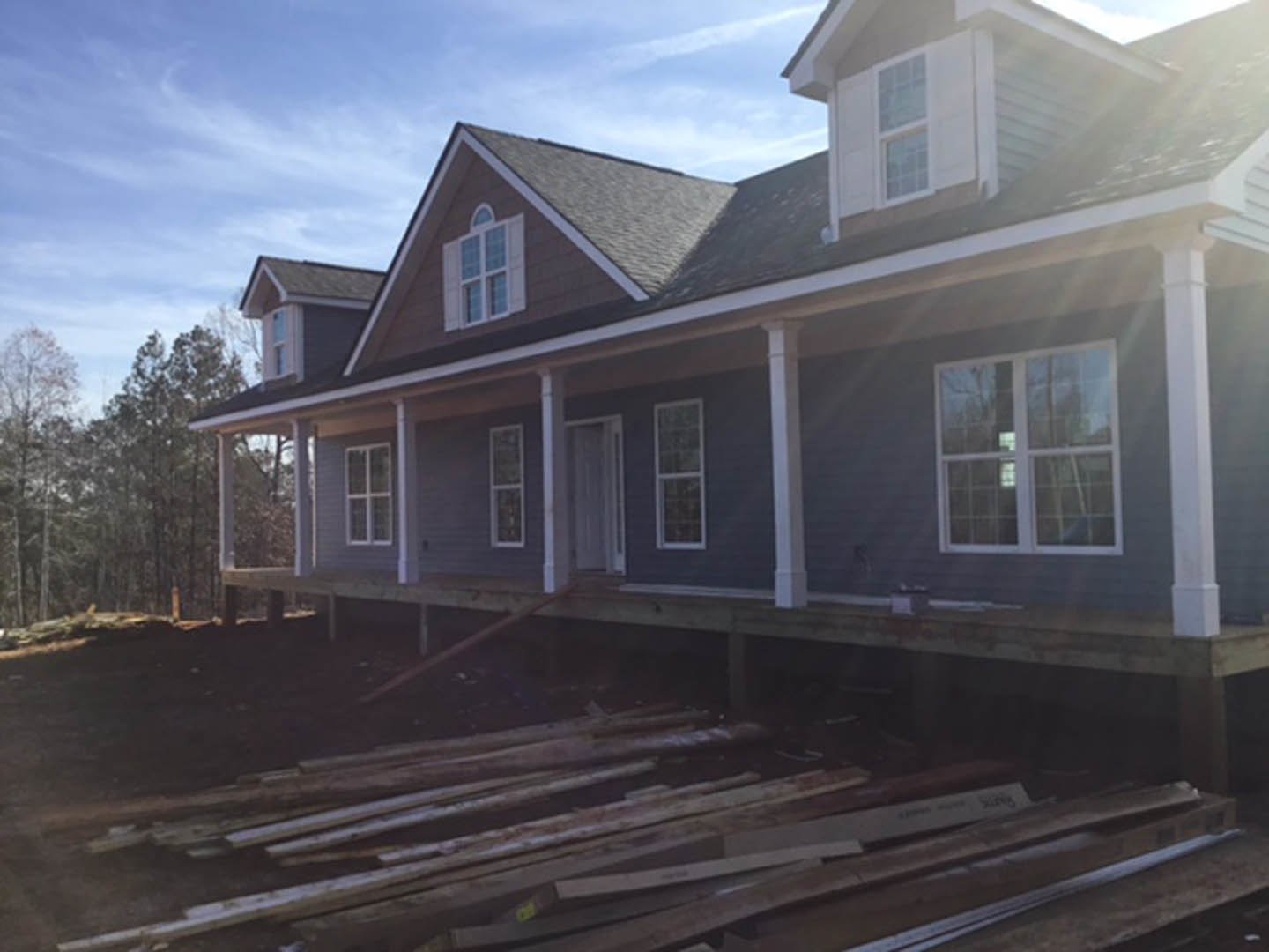 Framed house under construction with white window trim, exposed wood, and sunlight illuminating unfinished porch; trees and blue sky in background