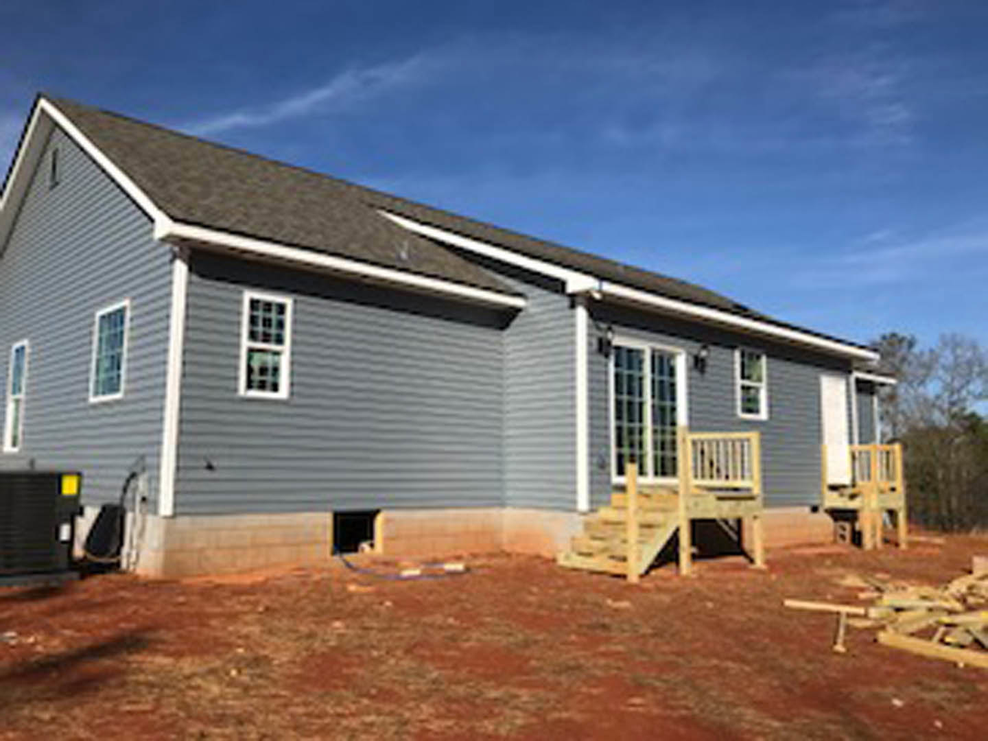 Wood-framed house under construction with unfinished wooden deck and railing, white window frames, gray siding, and clear blue sky in background