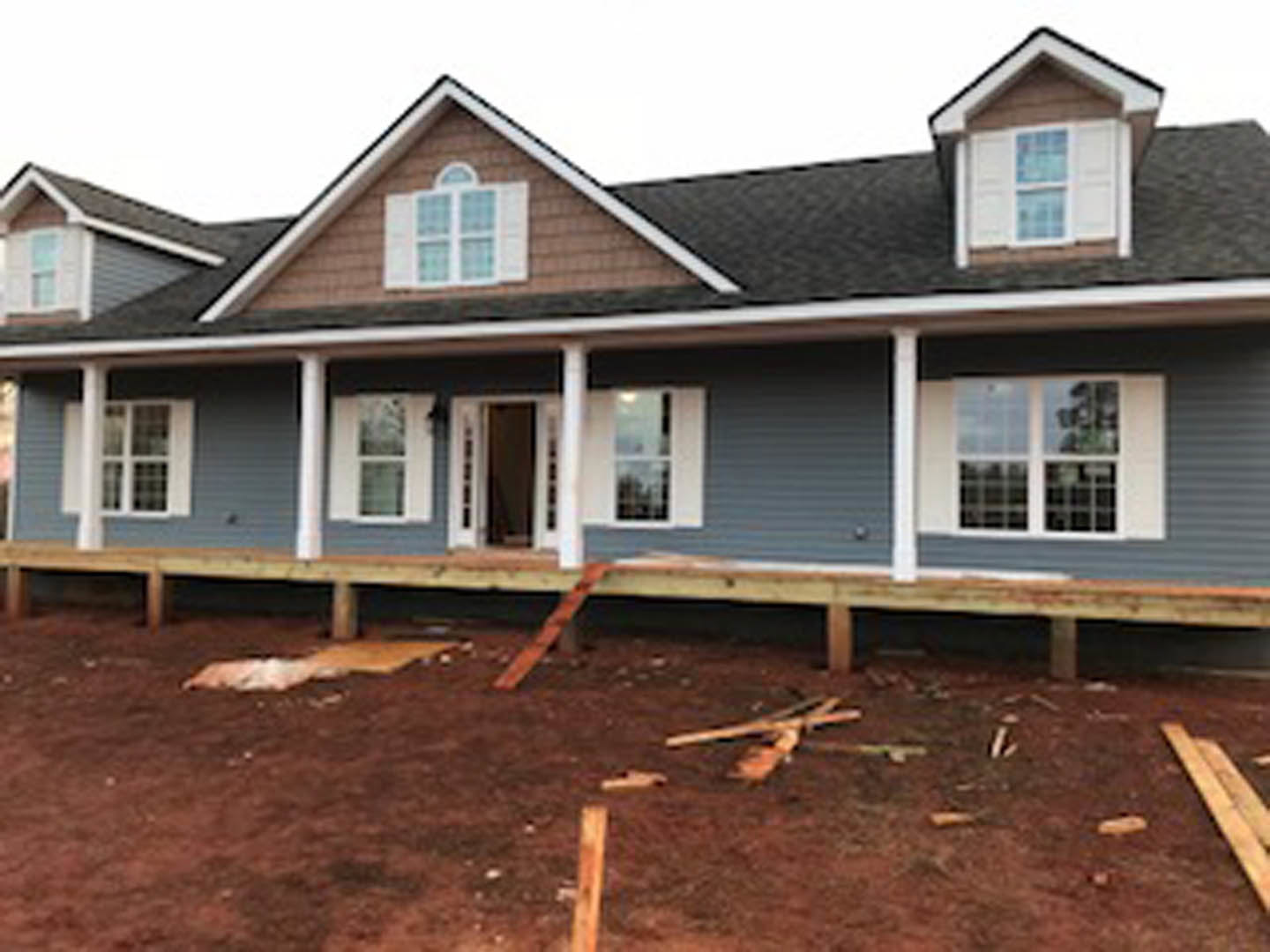 Two-story home under construction with exposed framing, unfinished porch, red dirt yard, scattered wood pieces, and visible windows and siding.
