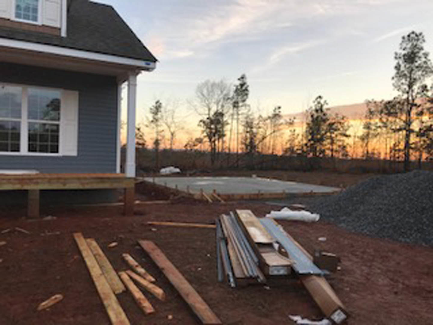 Modern home exterior with light siding, large windows, and a pile of lumber and gravel on the ground near the property, surrounded by trees and cloudy sky