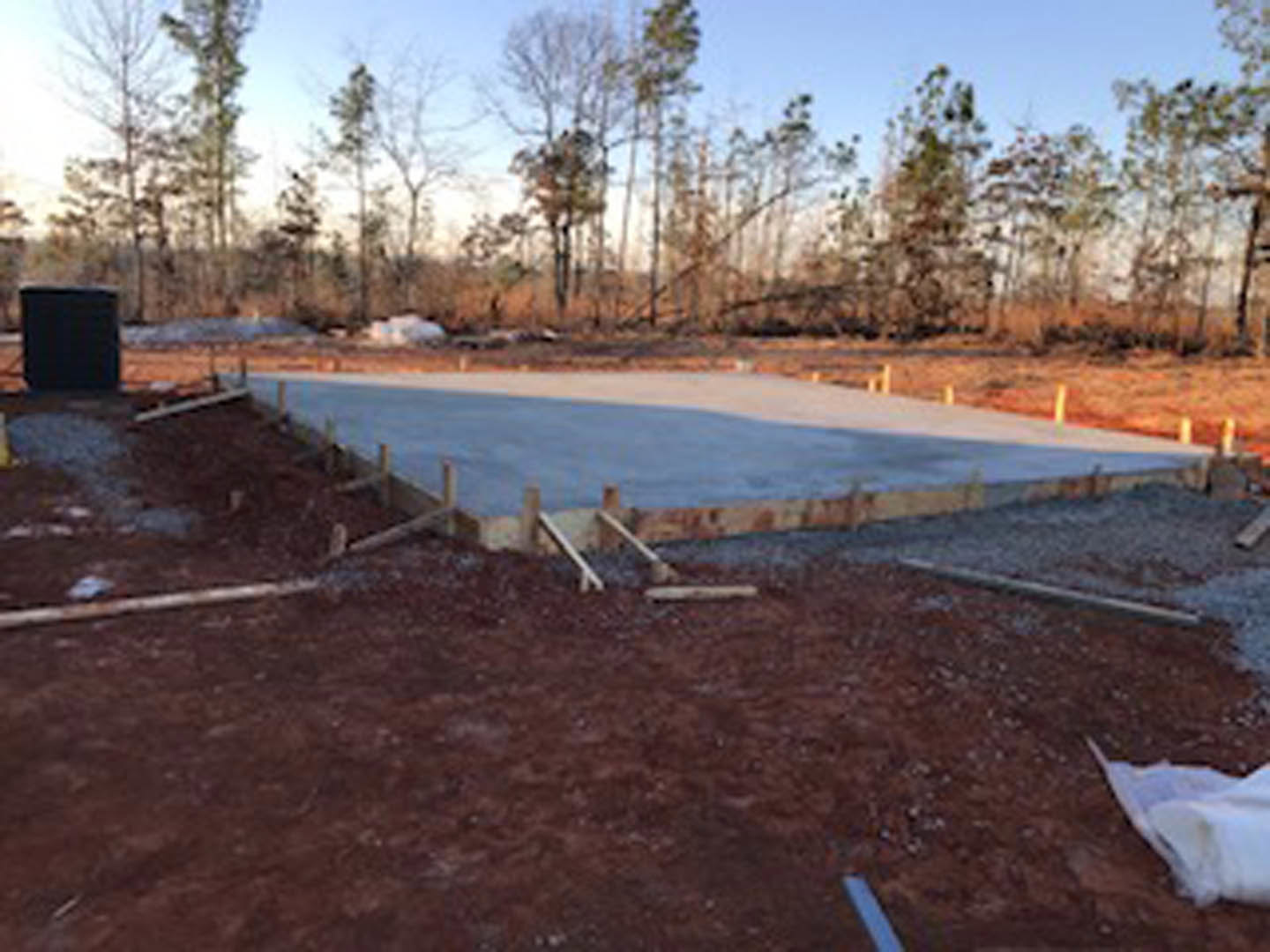 Concrete slab foundation under construction with wooden beams, dirt-filled site, scattered white bag, trees in background, and a man standing on a bench.