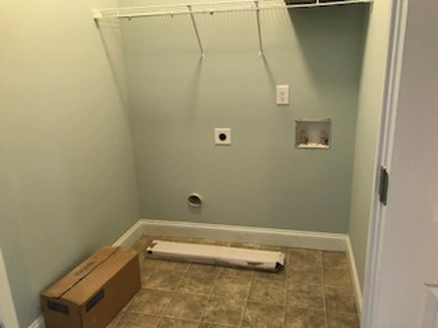 White plaster wall with a small hole, brown cardboard box on tile floor, minimal bathroom interior with visible ceiling and flooring.