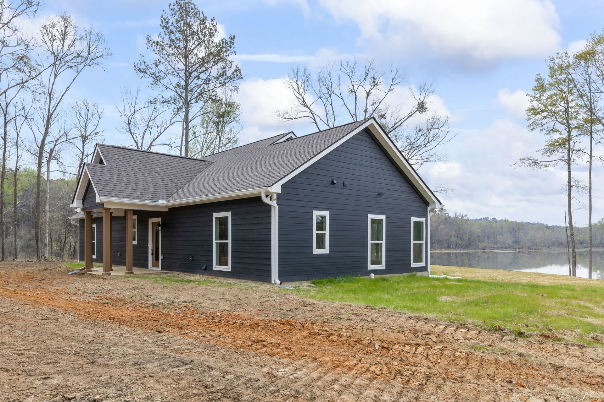 White-roofed house with white-framed windows, surrounded by grass and trees, dirt patch in front, lake visible in the background under a cloudy sky