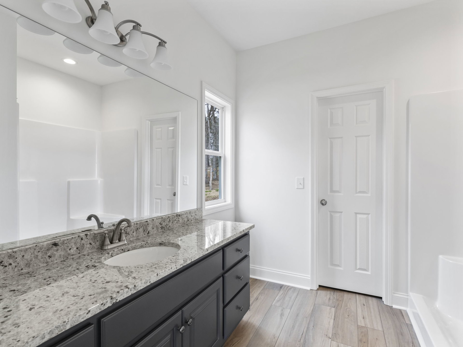Marble countertop with undermount sink, white cabinetry, large framed mirror, silver faucet, white door with silver knob, window overlooking trees, light tile flooring