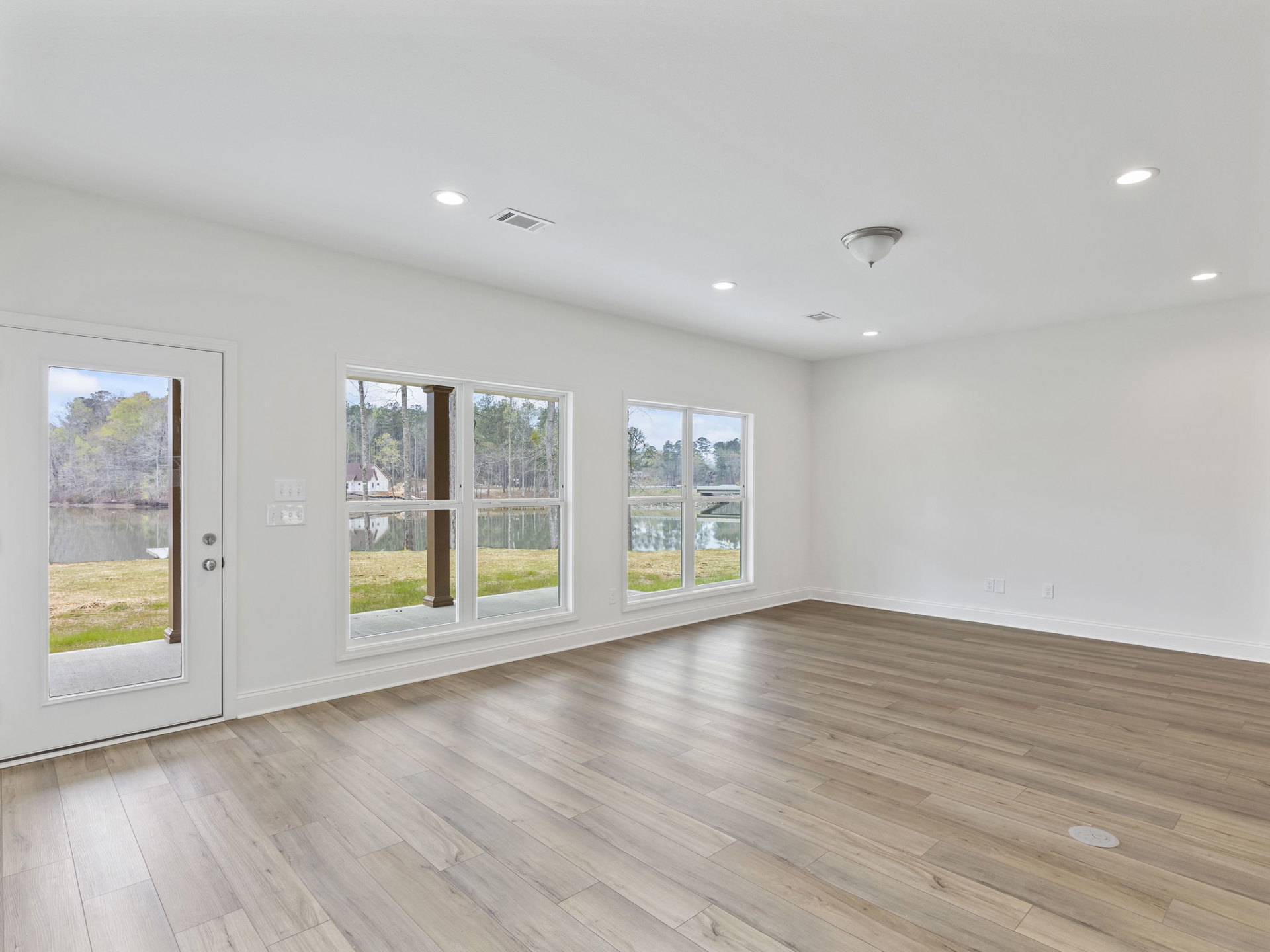 White walls and hardwood floors in a bright room, ceiling light fixture, large windows and door offering views of a lake and surrounding trees.