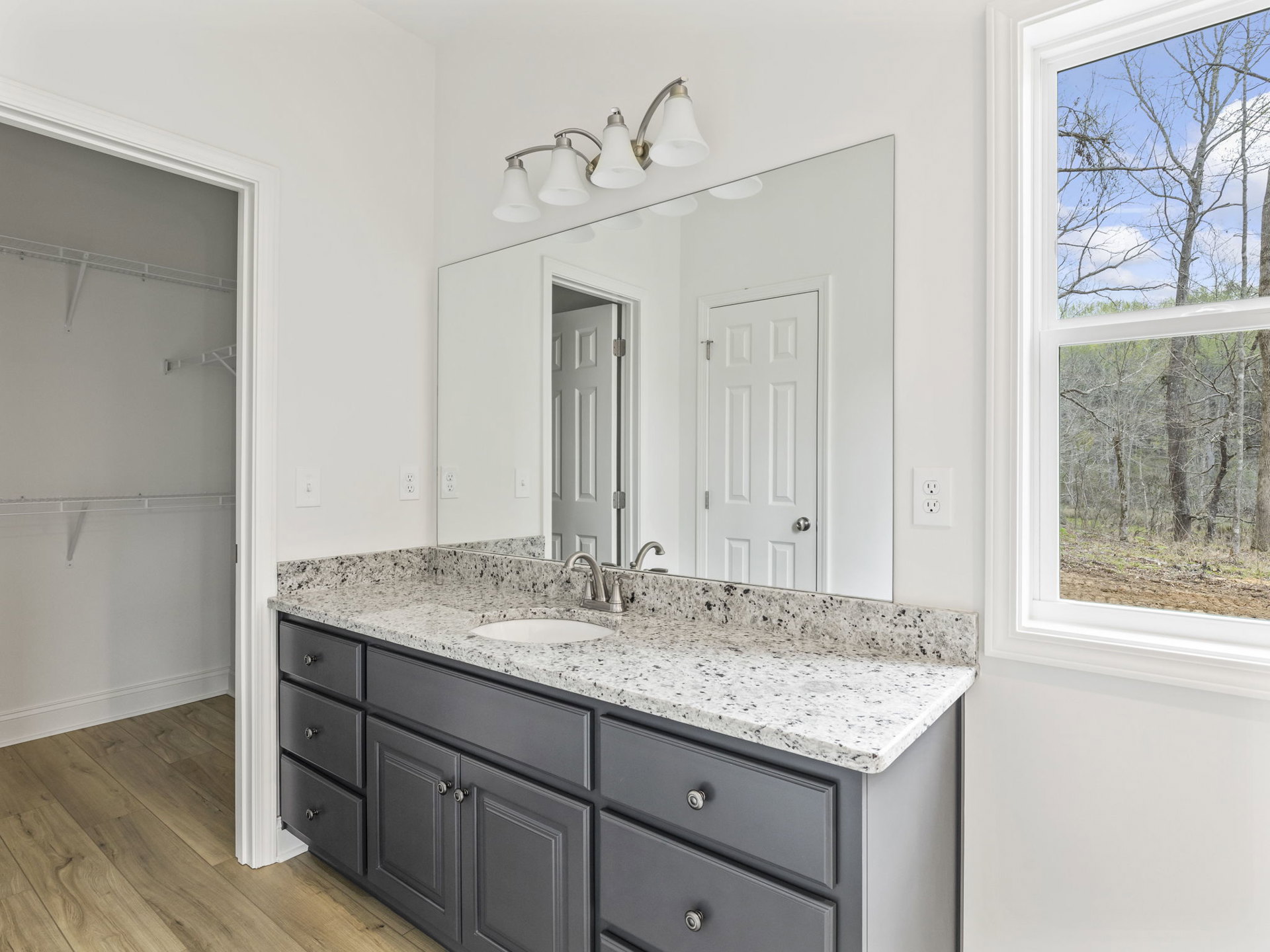 Bathroom featuring a wide mirror above a grey countertop sink, four white lamp light fixture, tile walls, cabinetry with drawers, and a window overlooking trees and sky.