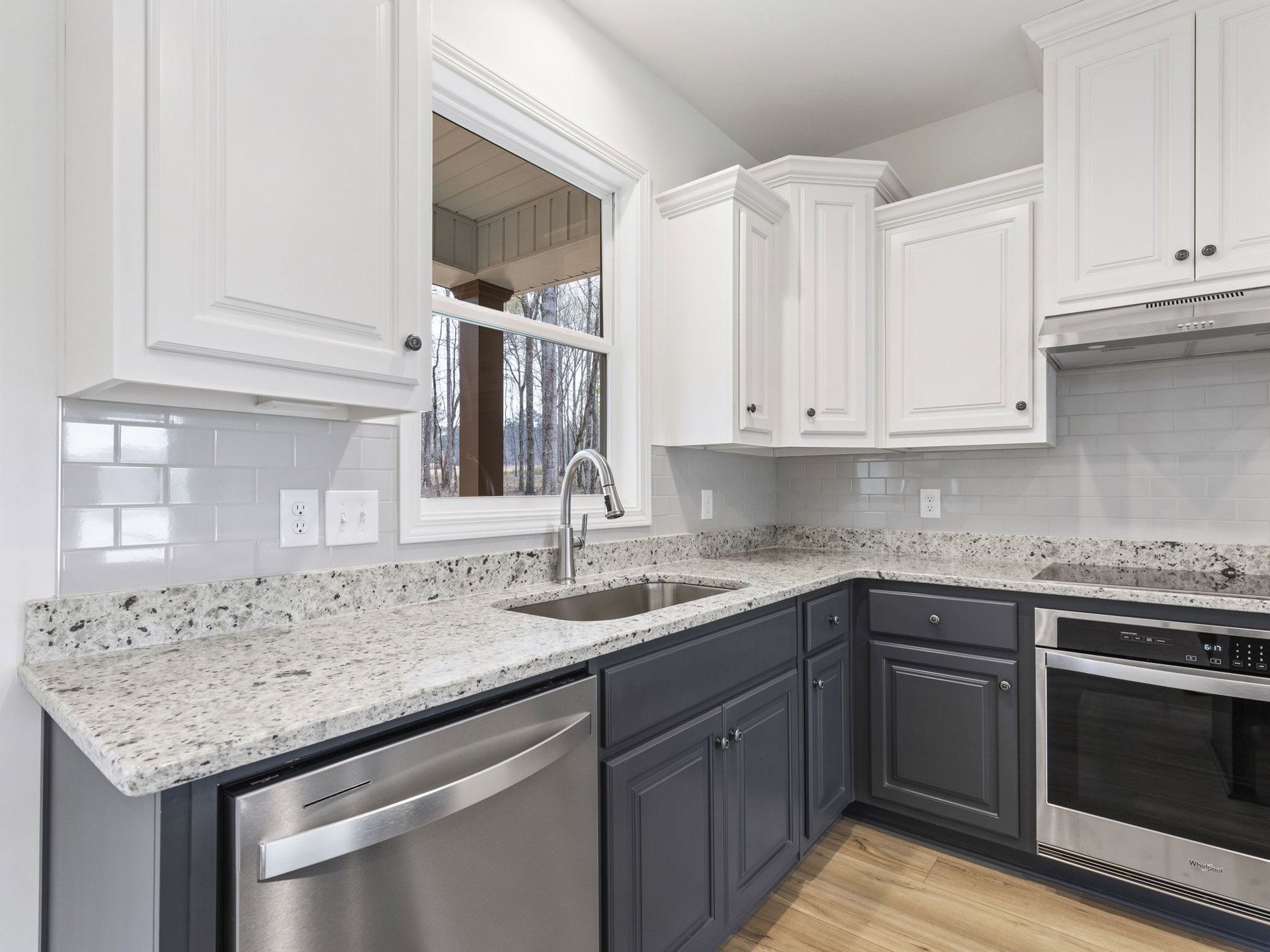 White kitchen cabinets and granite countertops with stainless steel microwave, dishwasher, and sink; window overlooks trees and pillar.