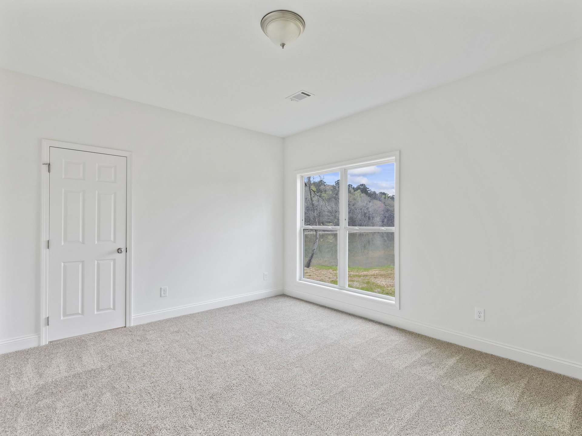 Carpeted bedroom with a white door, silver knob, large window overlooking trees, ceiling light fixture, and white walls with crown molding