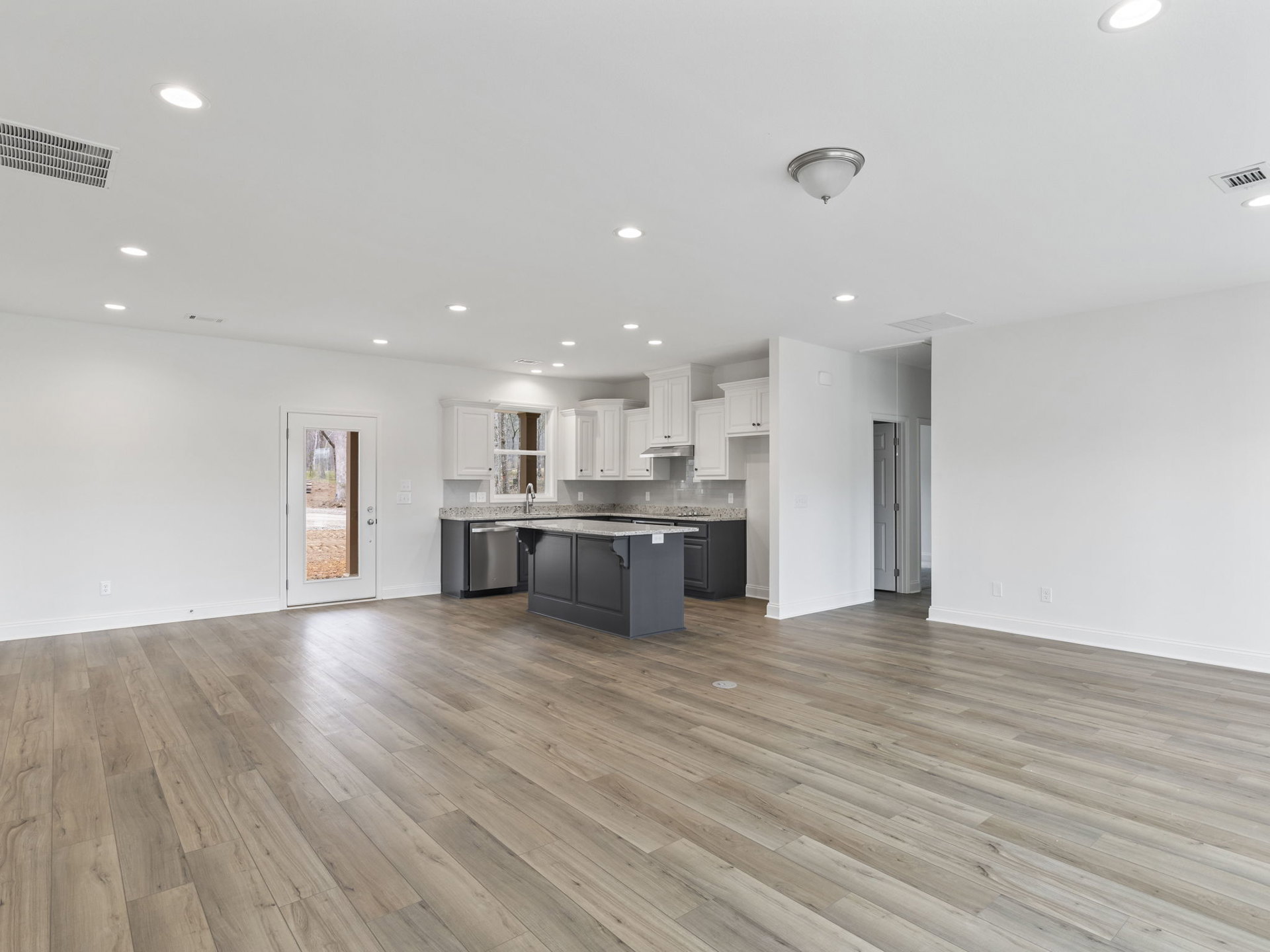 Spacious kitchen with marble-topped island, hardwood floors, white plaster walls, ceiling light fixture, and door overlooking forest.