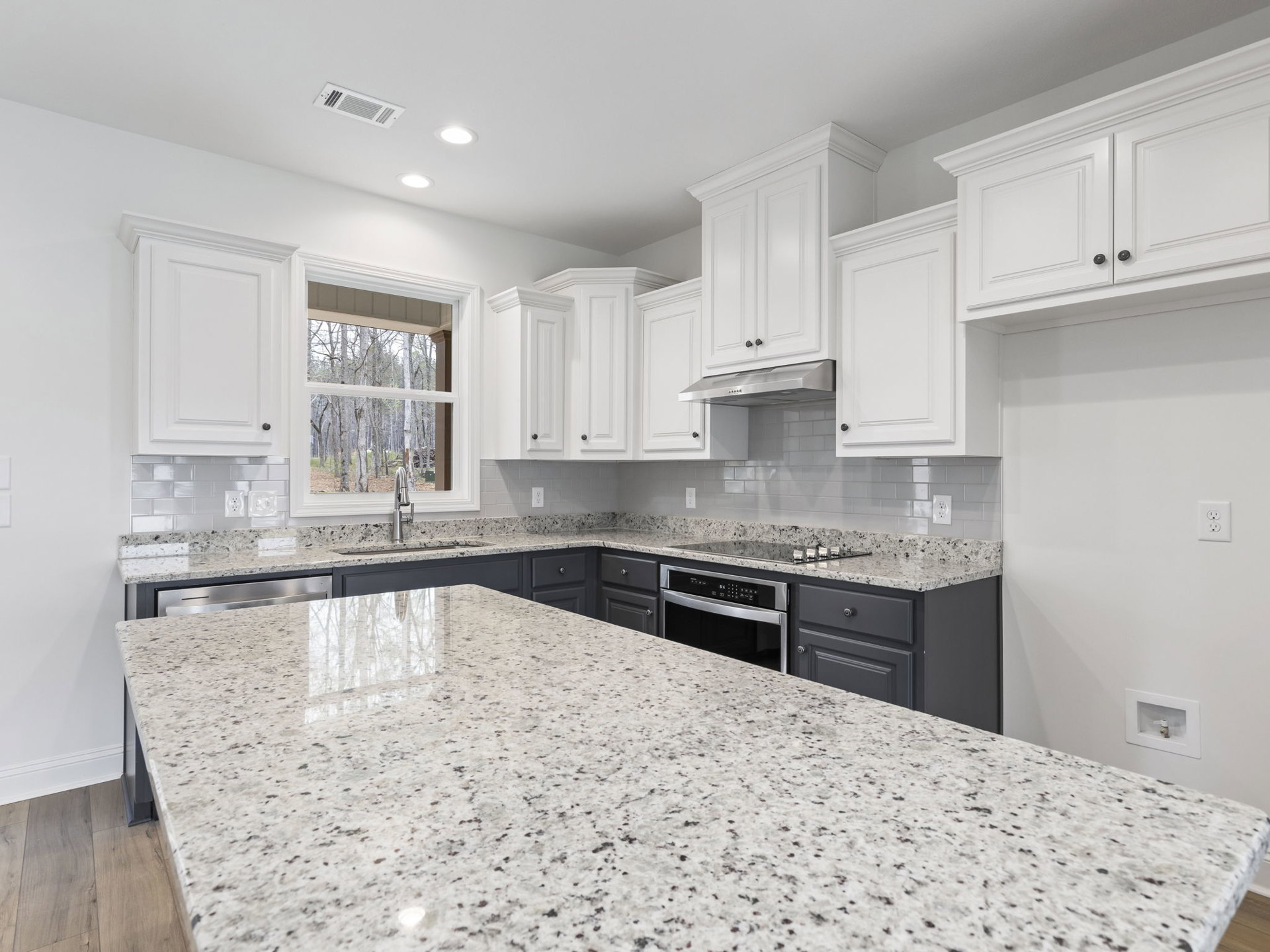 Granite countertops and white cabinets in a kitchen with stainless steel oven, built-in microwave, white ceiling vent, black-handled white door, and forest view through window
