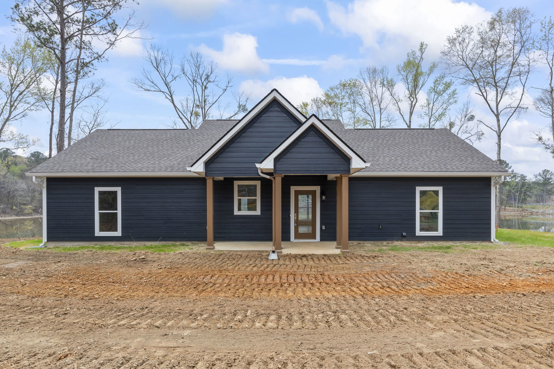 Two-story house under construction with blue roof, white-framed windows, glass-panel door, dirt patch with tire tracks, surrounded by trees and cloudy sky