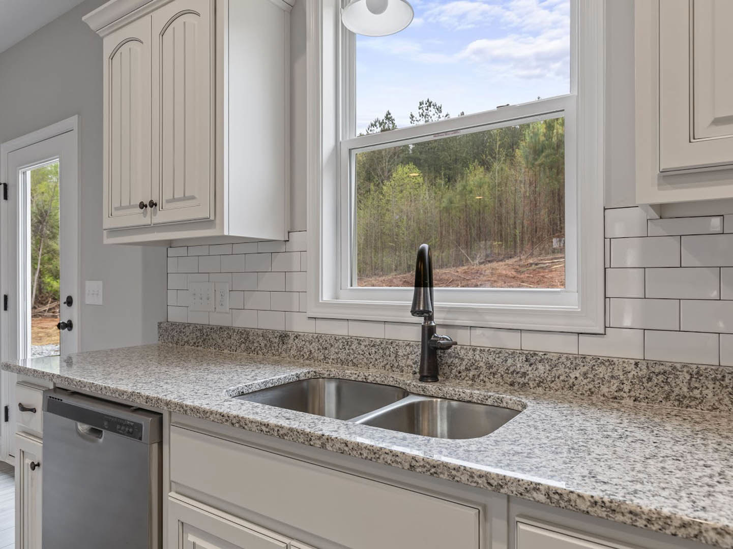 Modern kitchen with stainless steel sink and chrome faucet beneath a window overlooking trees, white cabinetry, built-in dishwasher, and paper towel roll on the countertop