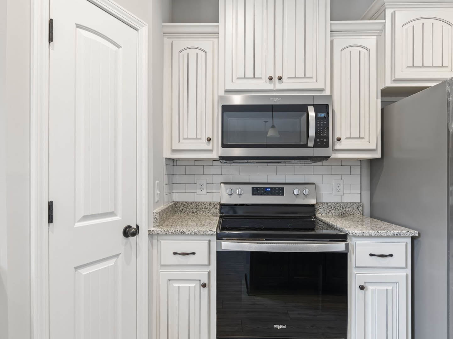 White kitchen cabinets, stainless steel microwave above a black glass stove, white countertop, black door knob on adjacent white door, white lamp shade in background
