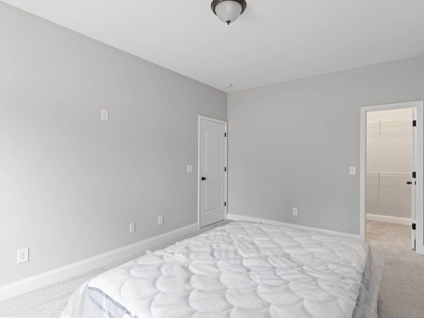 White mattress with crisp bedding placed directly on light wood floor, adjacent to white paneled door with black hardware, wall outlet visible near baseboard, minimal decor and