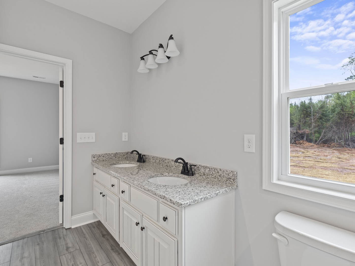 Bathroom with marble countertops, white toilet, wall-mounted light fixture, window overlooking trees, tile flooring, and cabinetry.