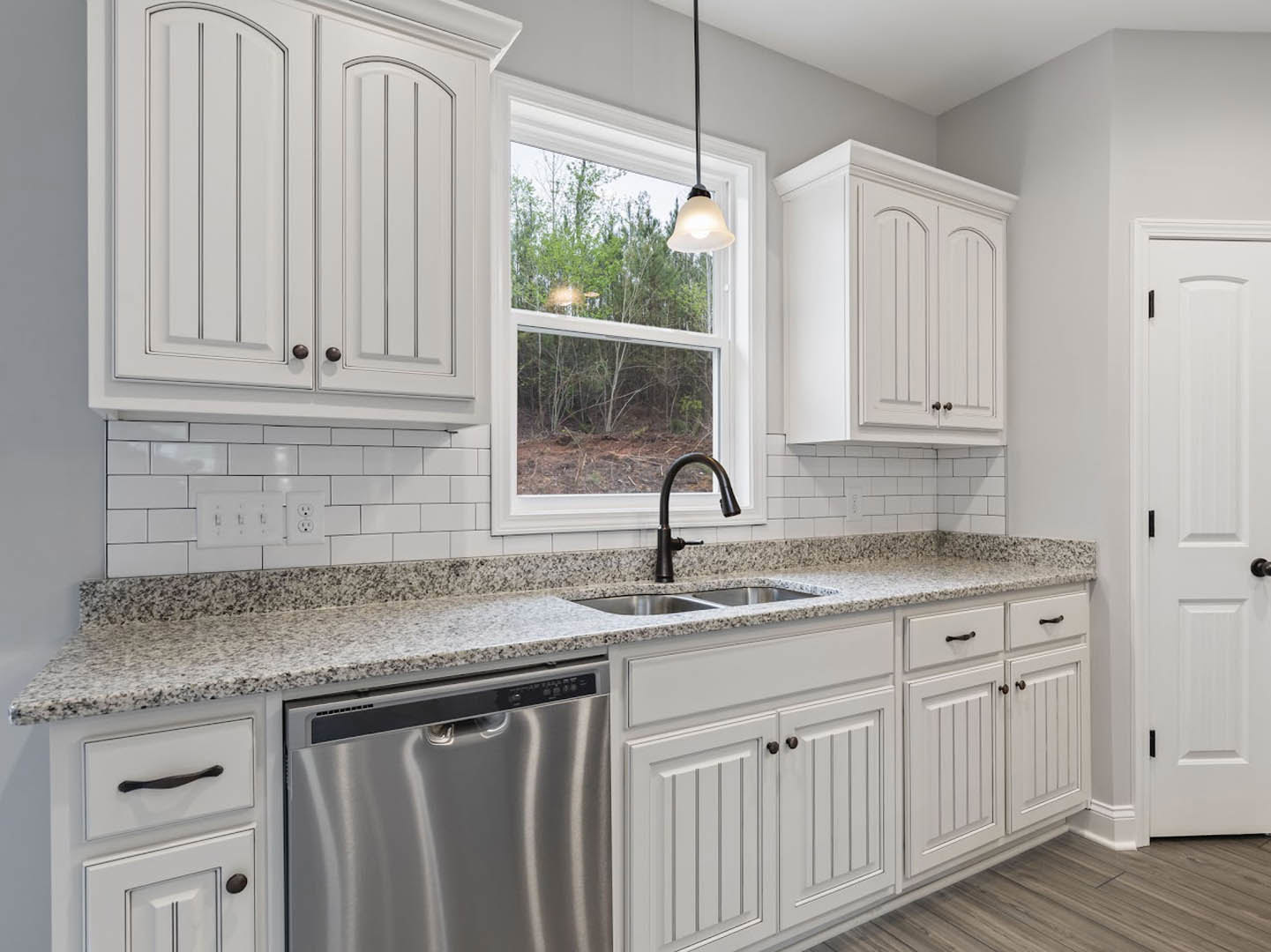 White kitchen cabinets, stainless steel sink with modern faucet, built-in dishwasher, quartz countertop, window overlooking trees, pendant light fixture, white door with black