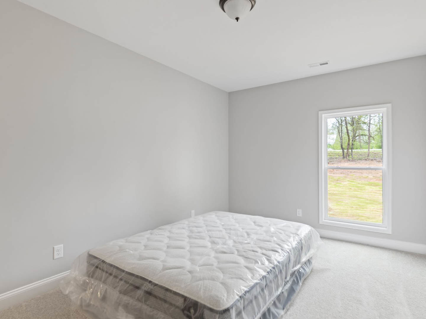 Queen bed with plastic-wrapped mattress and white linens, positioned near a window overlooking trees and a grassy field; white electrical outlet on wall, ceiling light fixture