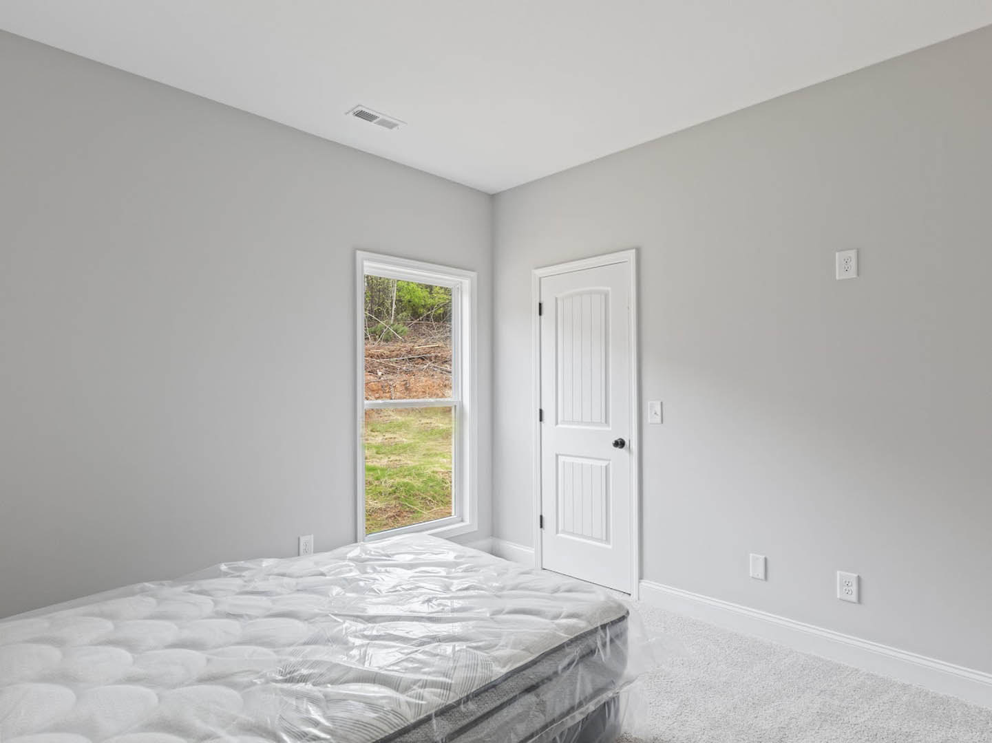 Queen bed with plastic-wrapped mattress, white walls, white door with black knob, large window overlooking grassy field, light wood flooring, white ceiling