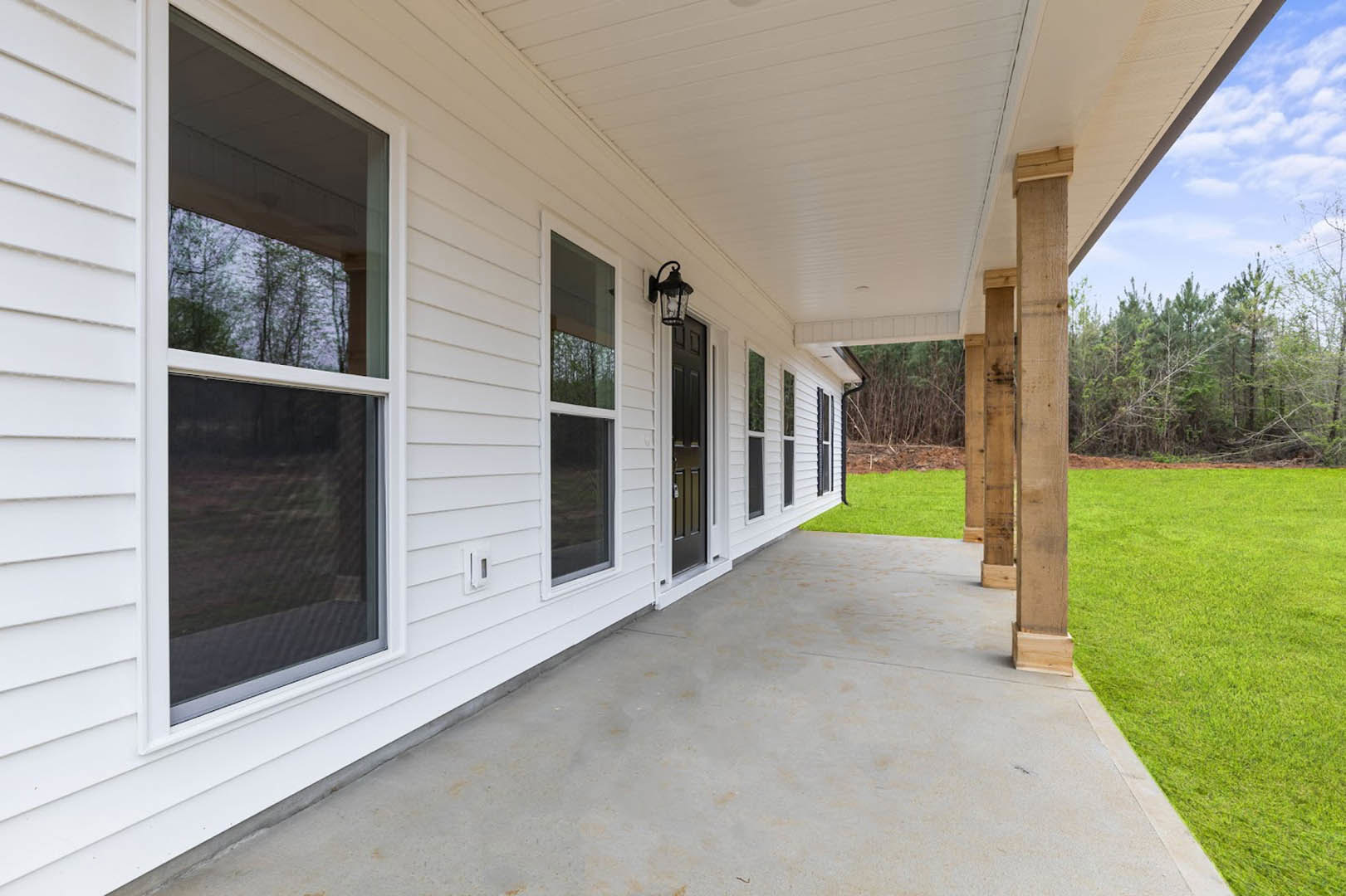 White siding house with covered front porch, black-framed windows, green grass lawn, outdoor lamp, and leafy trees visible through windows