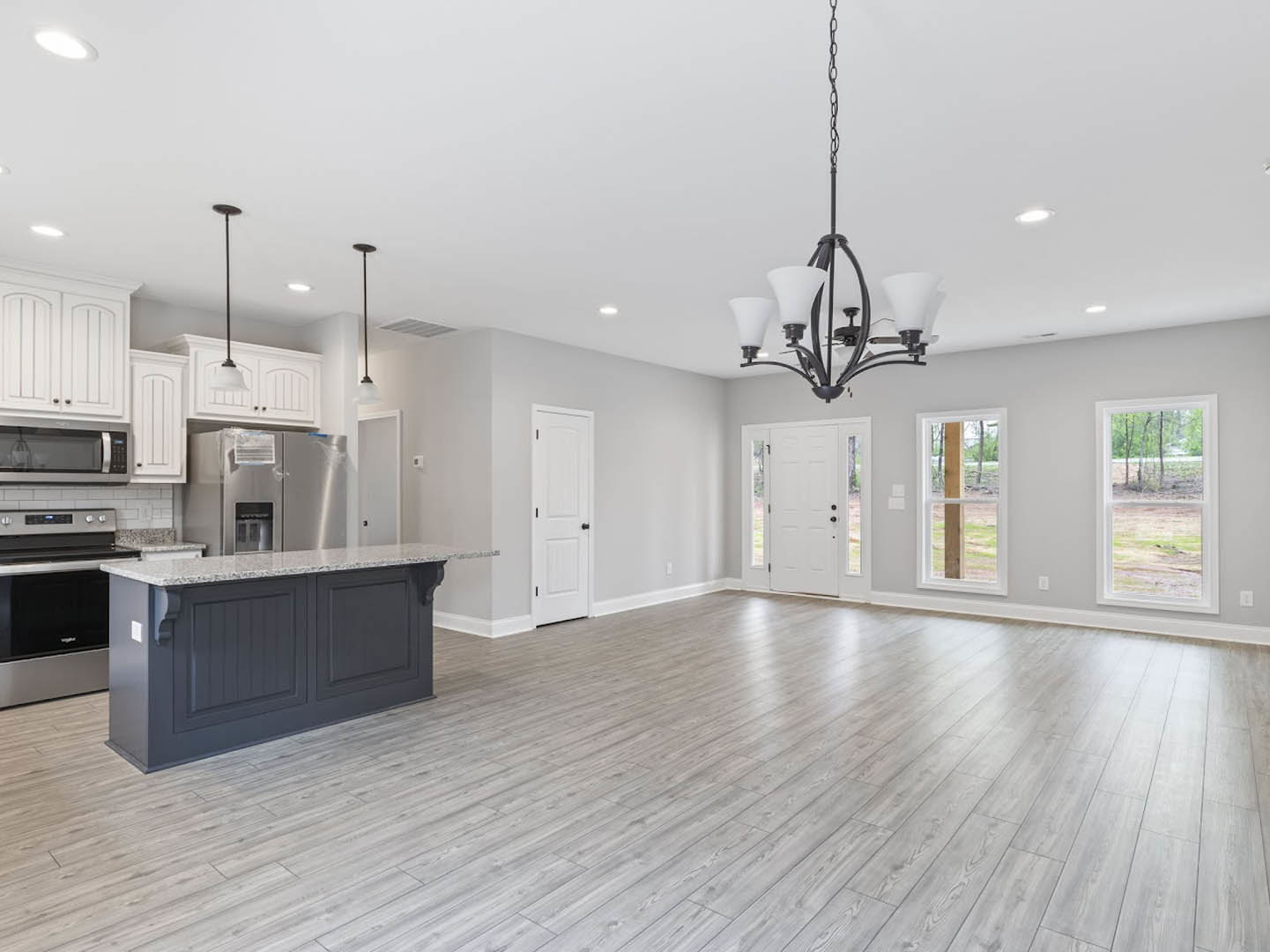 Spacious open-concept kitchen with wood flooring, central island, white cabinetry, black microwave, large window overlooking field and trees, modern chandelier with white shades