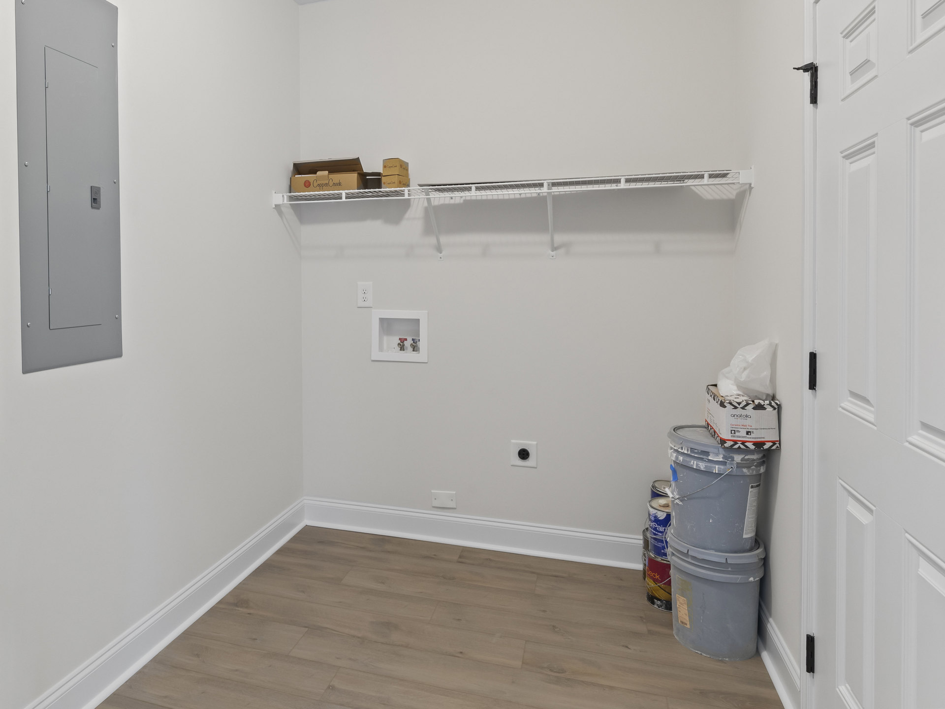 White-walled room with built-in shelves, grey door featuring a hole, wood flooring with white baseboard, white utility box with two valves, and grey bucket topped with white paint.
