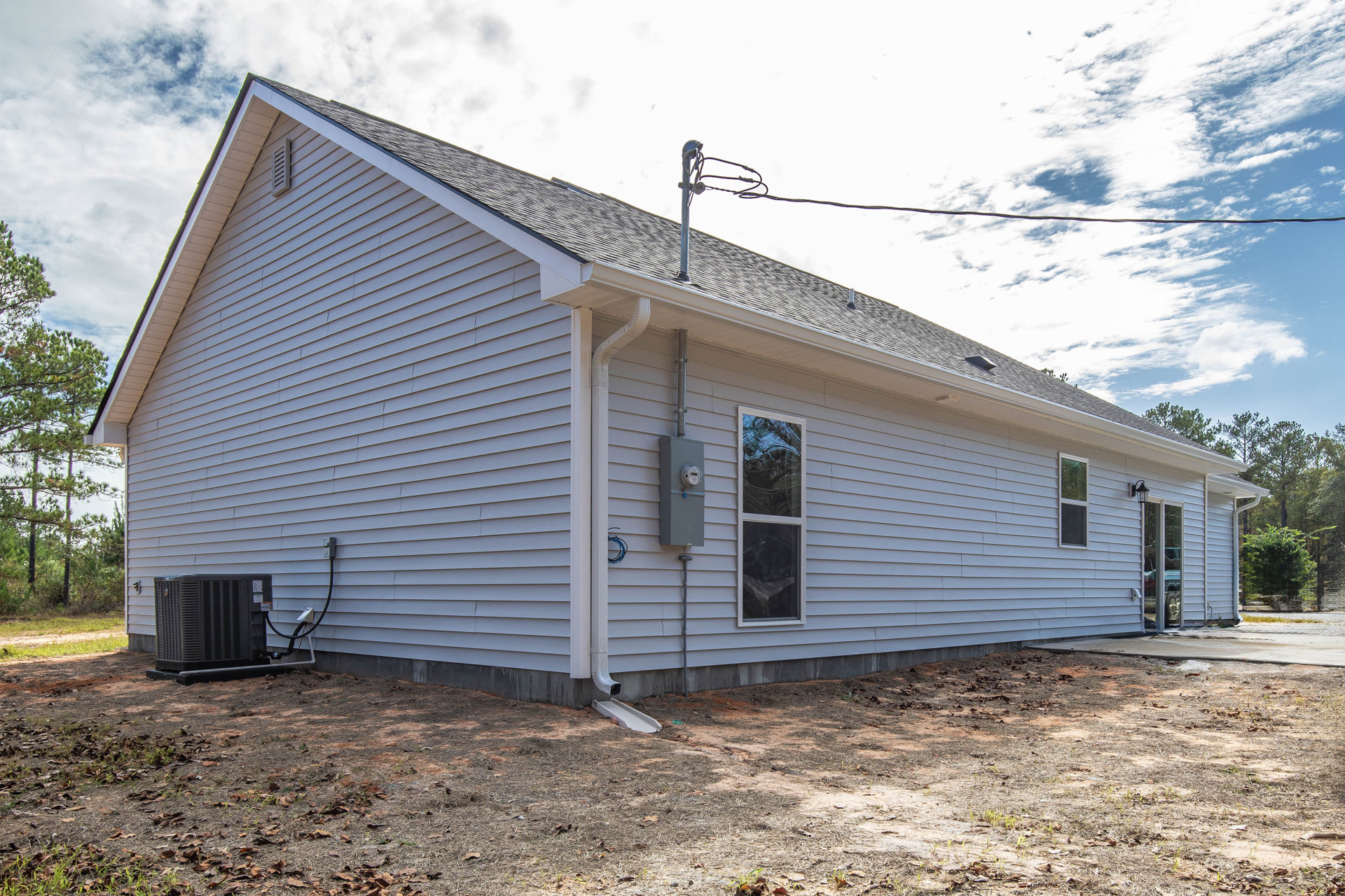 Modern house exterior with light-colored siding, large window reflecting nearby trees, black heat pump unit, gutter along the roof edge, electrical meter mounted on the wall