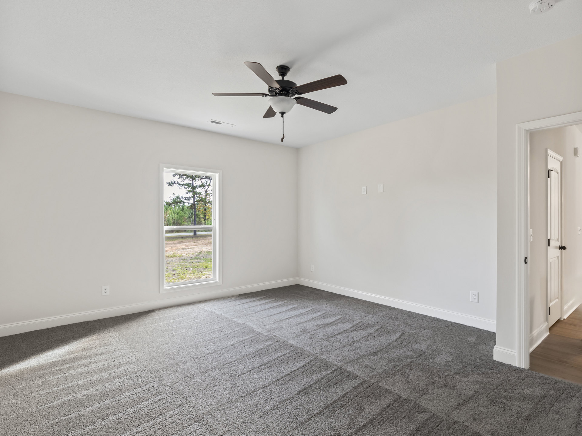 Ceiling fan with light fixture above grey carpeted floor, white walls, large window showing trees and grass outside