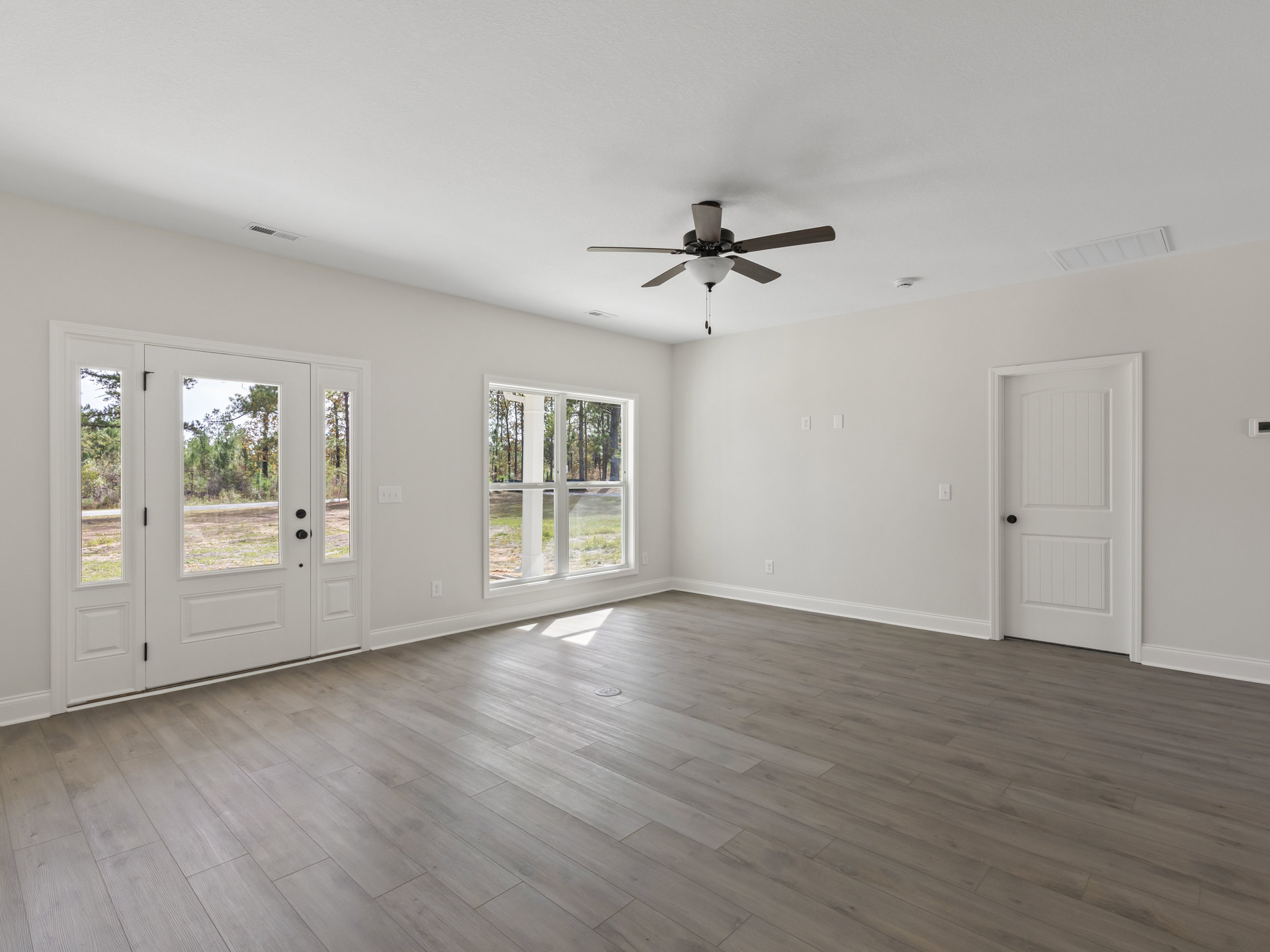 White-walled room with wood flooring, ceiling fan with light fixture, window overlooking trees, and white door with black handle