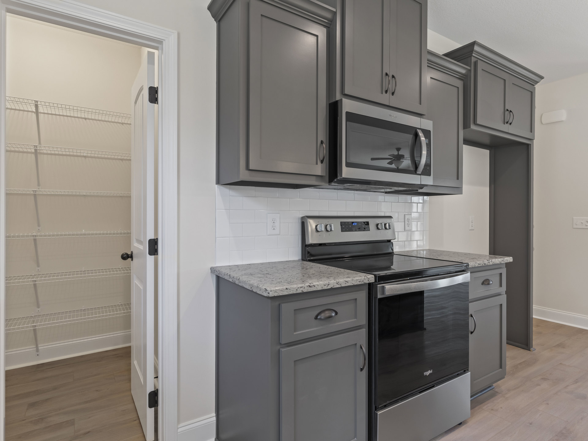 Grey shaker cabinets, stainless steel microwave with vent hood, white door with black handle, light quartz countertops, neutral tile backsplash