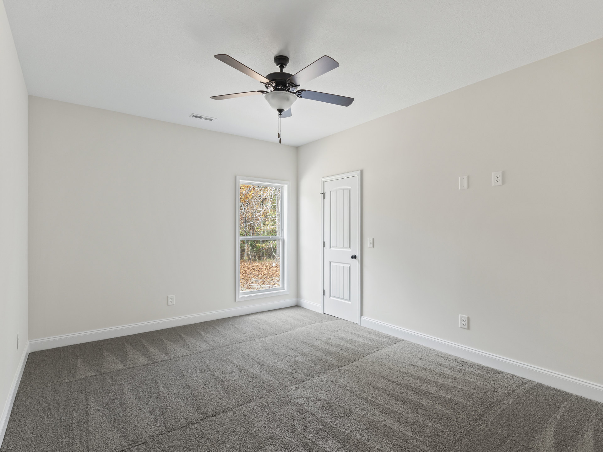 Carpeted room with white walls, ceiling fan and light fixture, white door with black knob, large window overlooking trees and forest.
