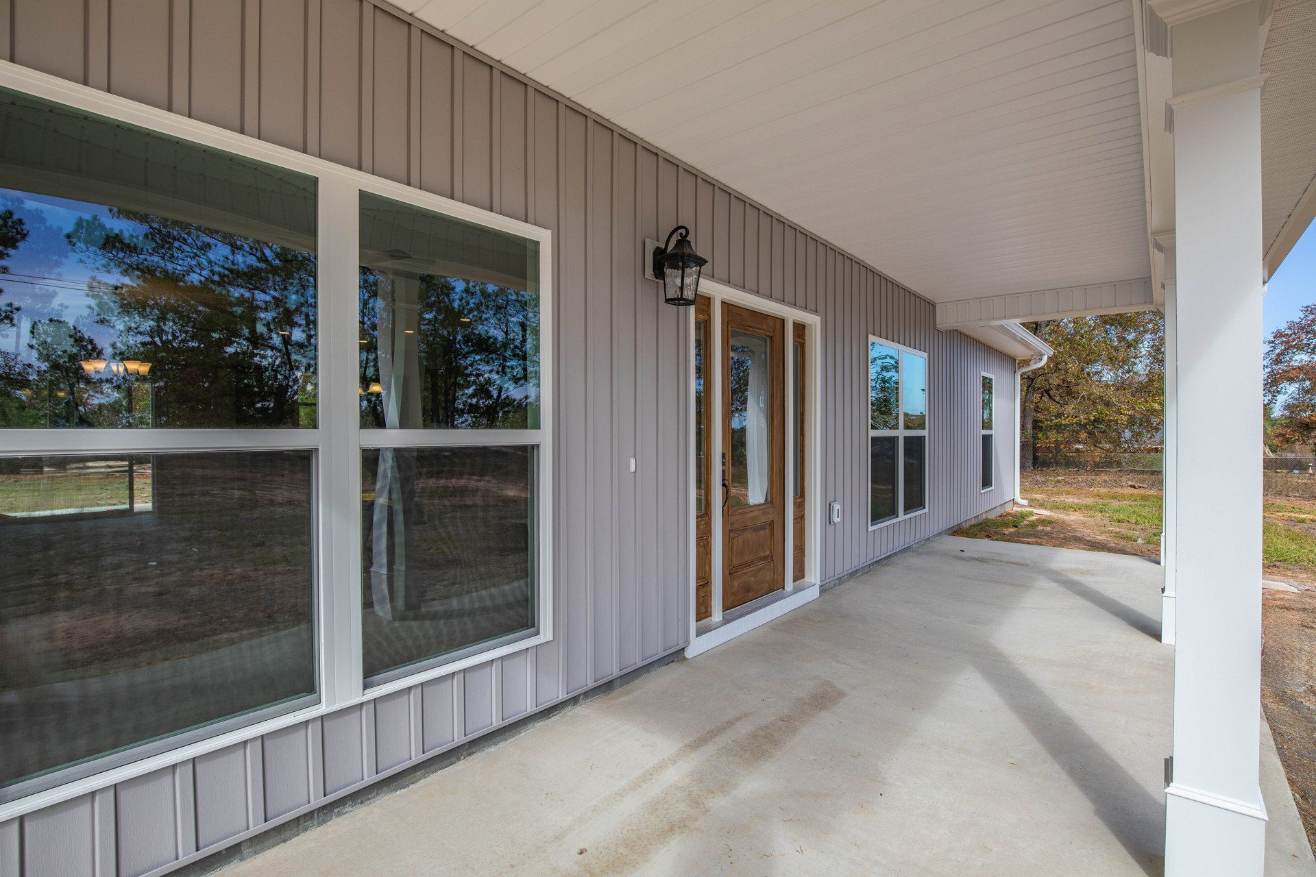White front porch with composite decking, glass-paneled door, large windows reflecting trees, and outdoor lamp fixture.
