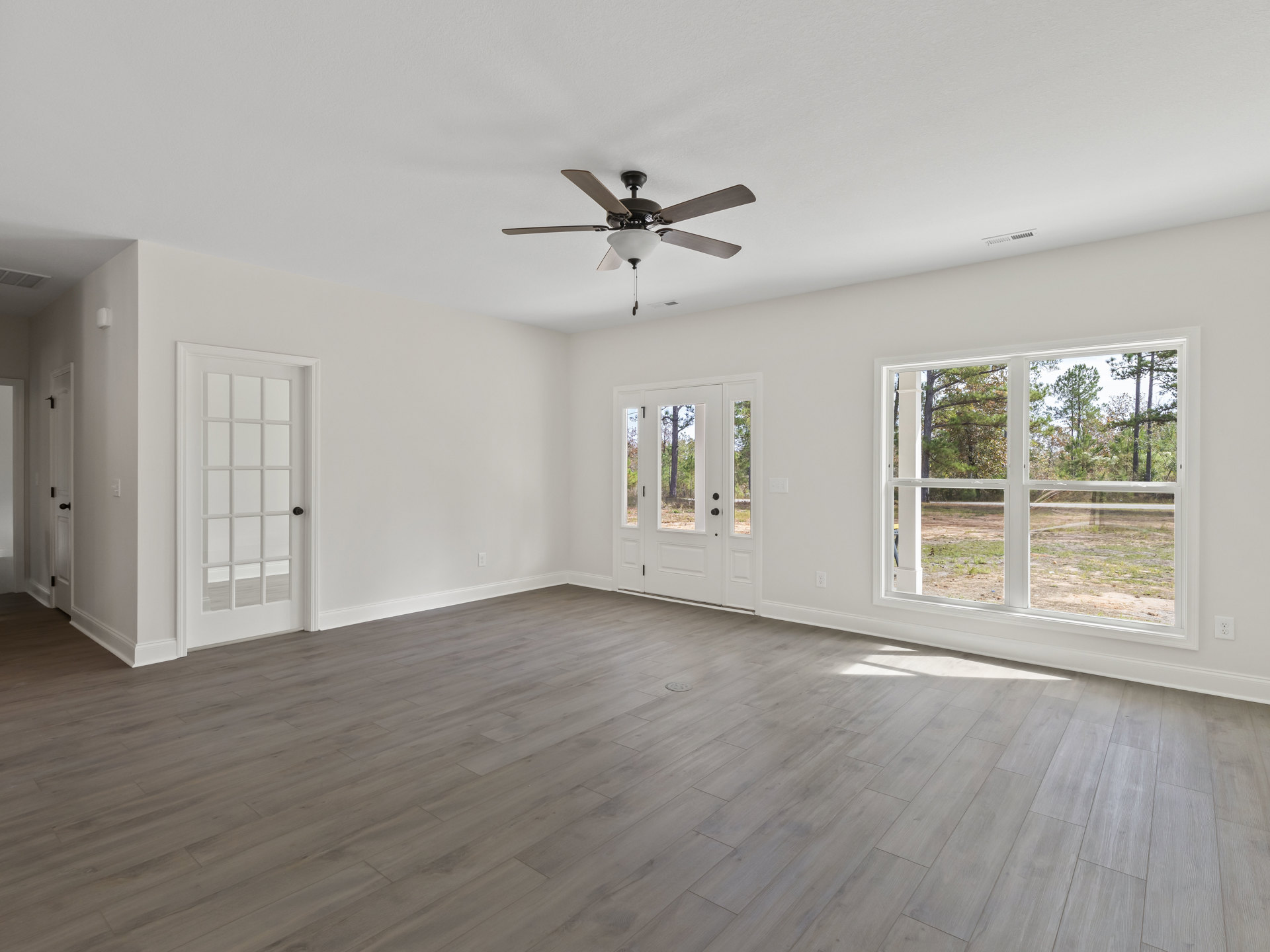 Ceiling fan with light fixture, wood laminate flooring, white door with glass panels, large windows showing trees outside, plaster walls and ceiling