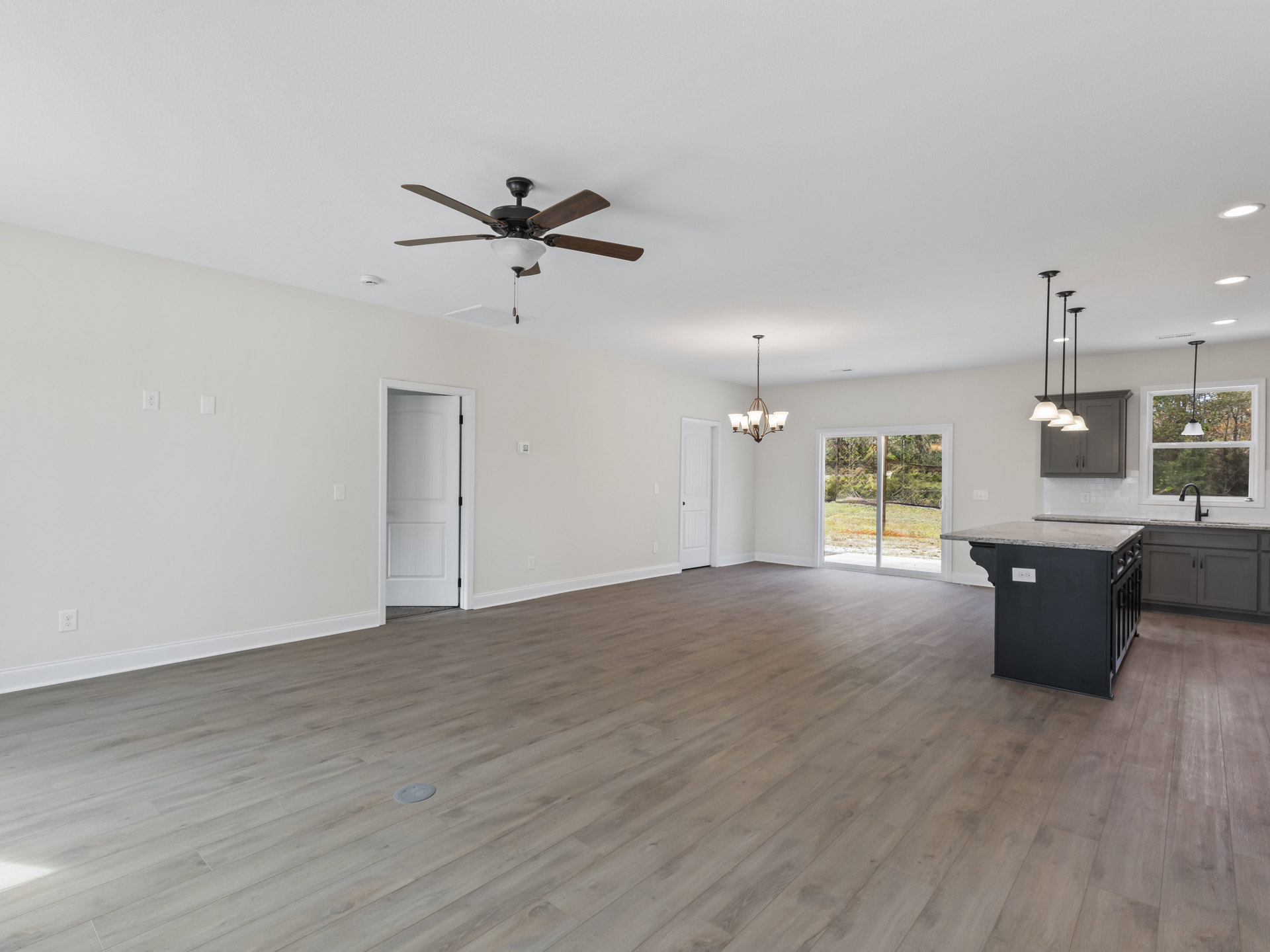 Spacious open-plan room featuring a marble-topped kitchen island, hardwood floors, white walls, ceiling fan with light, white door with black handle, and sliding glass door