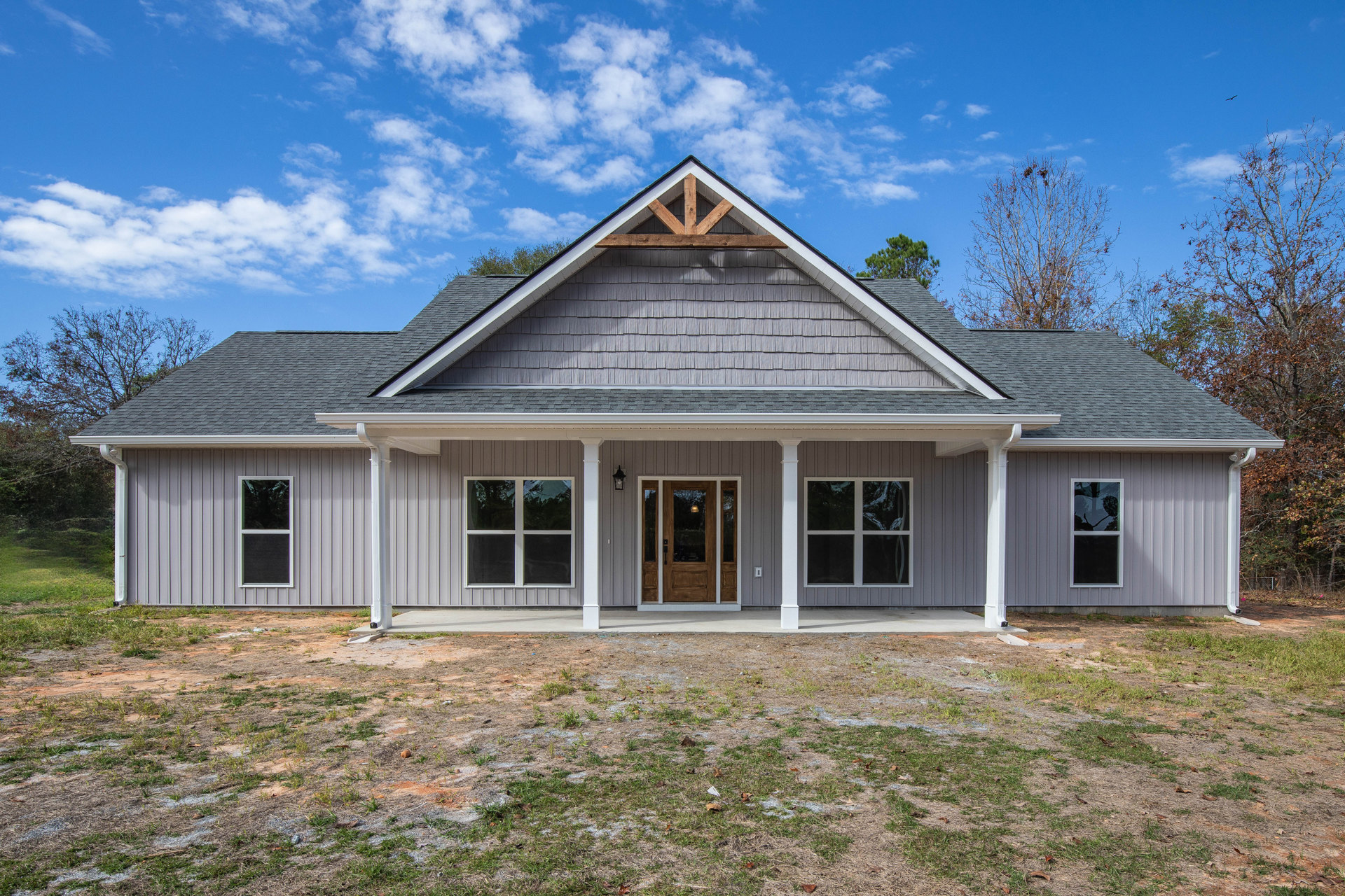 Two-story home with wood roof, brown front door with exterior light, white-framed windows reflecting trees, landscaped yard and mature trees in background