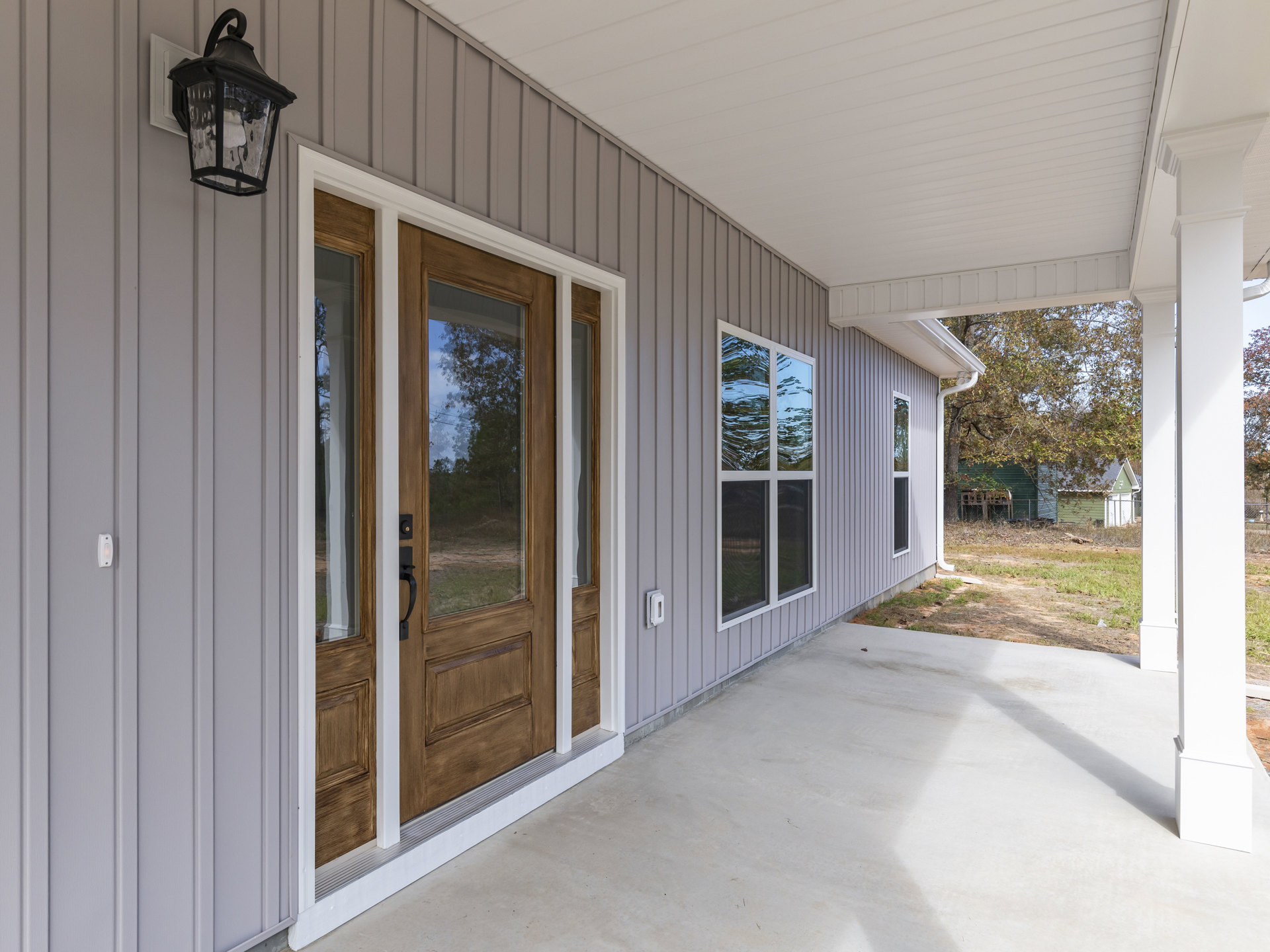 Front porch with concrete patio, glass-paneled door with metal handle, window featuring tree reflections and screen, black lantern fixture mounted beside entry.