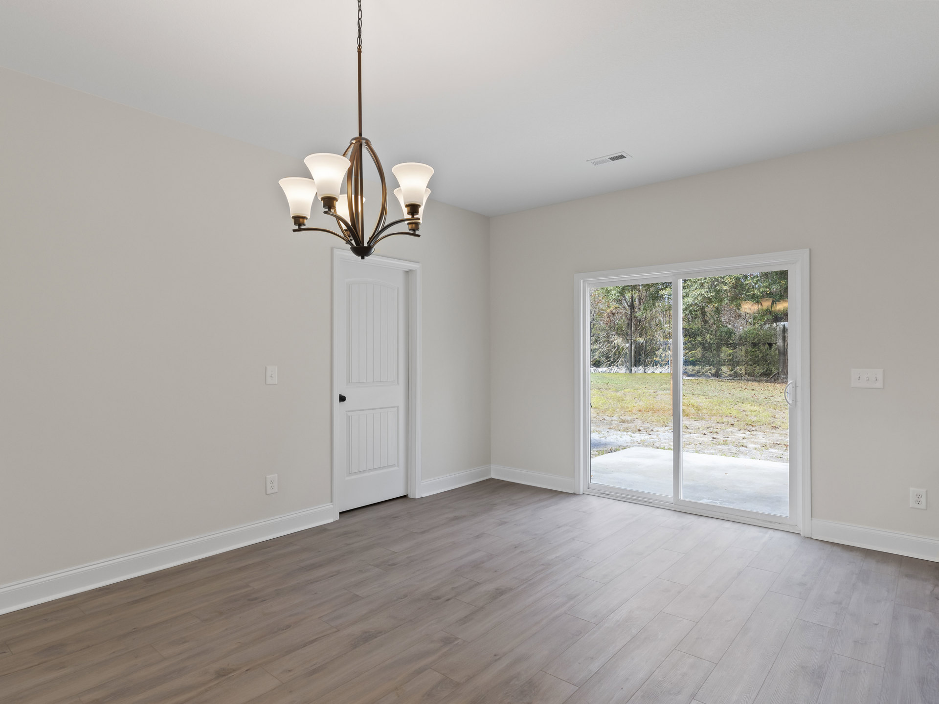 Wood flooring and white walls in a bright room with a sliding glass door overlooking a grassy field, modern chandelier hanging from the ceiling, and a white door with a black knob.